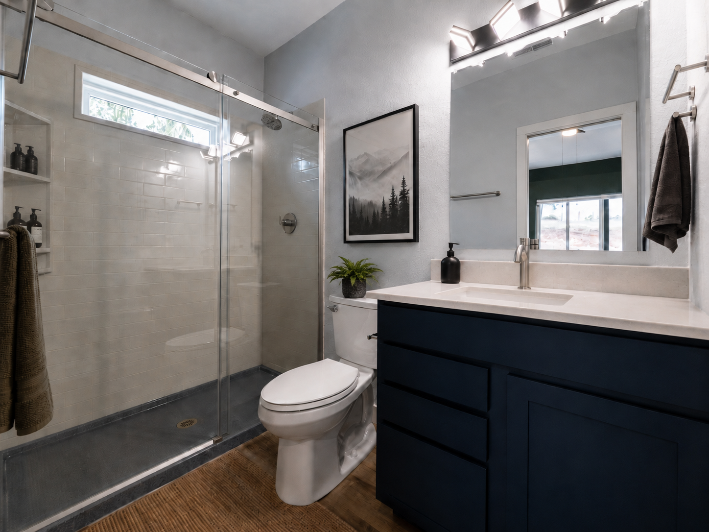 Modern bathroom with a glass shower enclosure, white subway tiles, a white toilet, a dark blue vanity with a light-colored countertop, a black soap dispenser, a small plant, a framed black-and-white mountain landscape photograph, a large mirror, and 