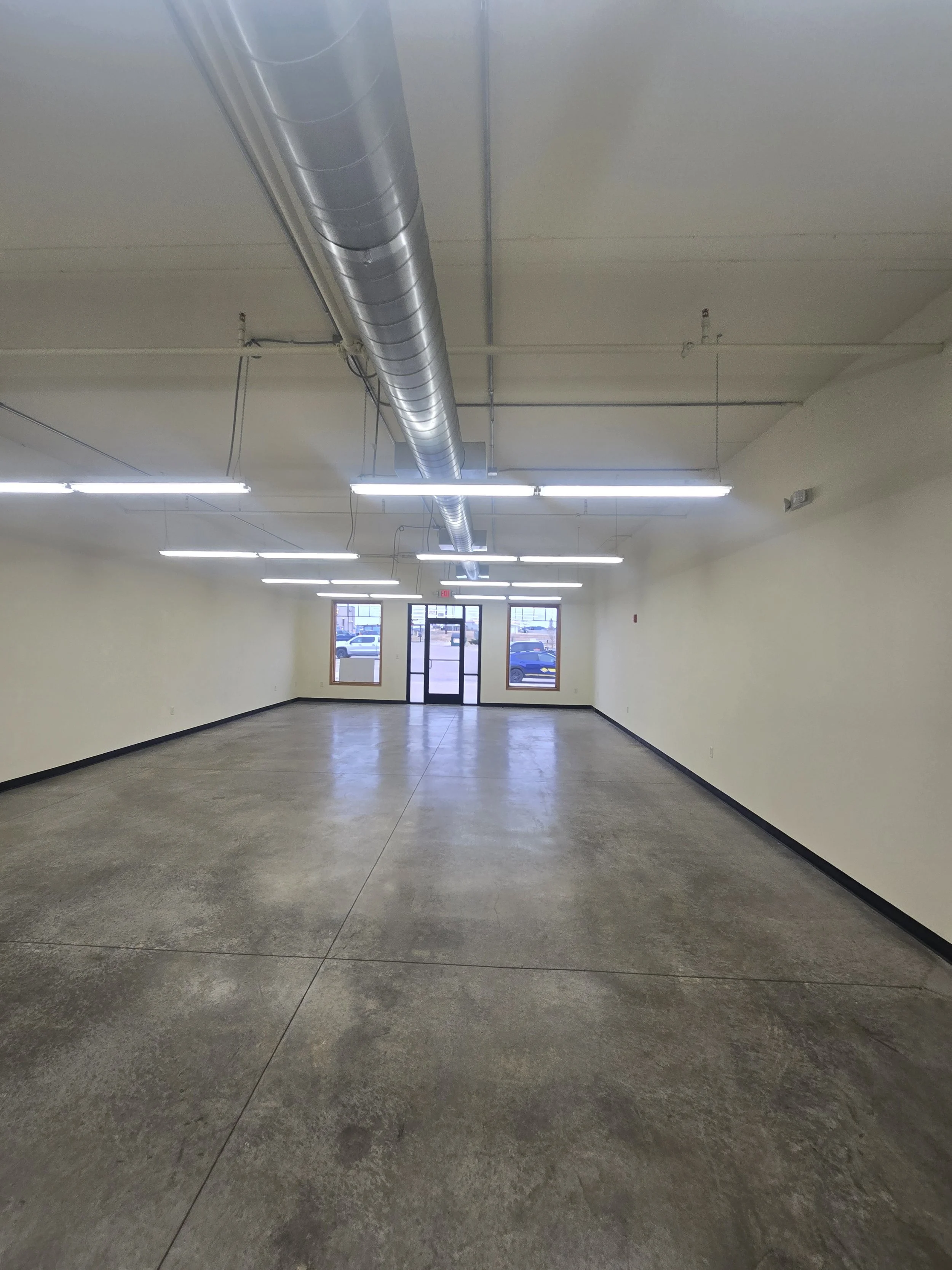Empty commercial space with concrete floor, white walls, ceiling-mounted fluorescent lights, large windows, glass door, and exposed ductwork.