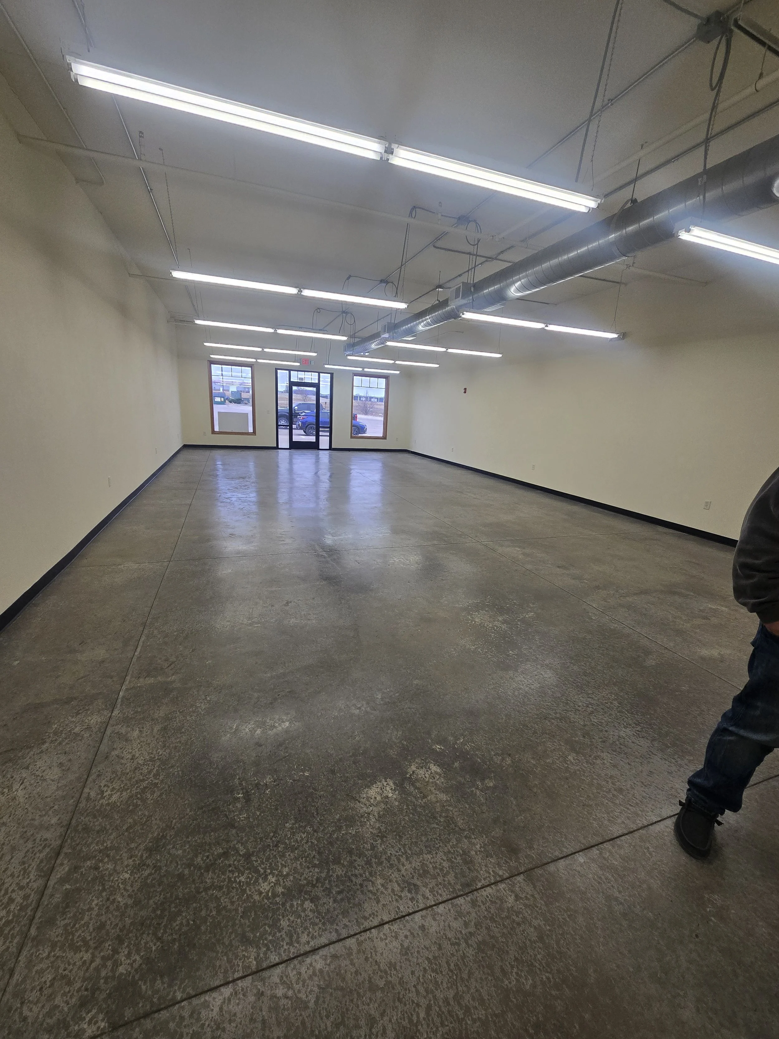 Empty commercial space with polished concrete floors, cream-colored walls, fluorescent lighting, and large front windows with a glass door.