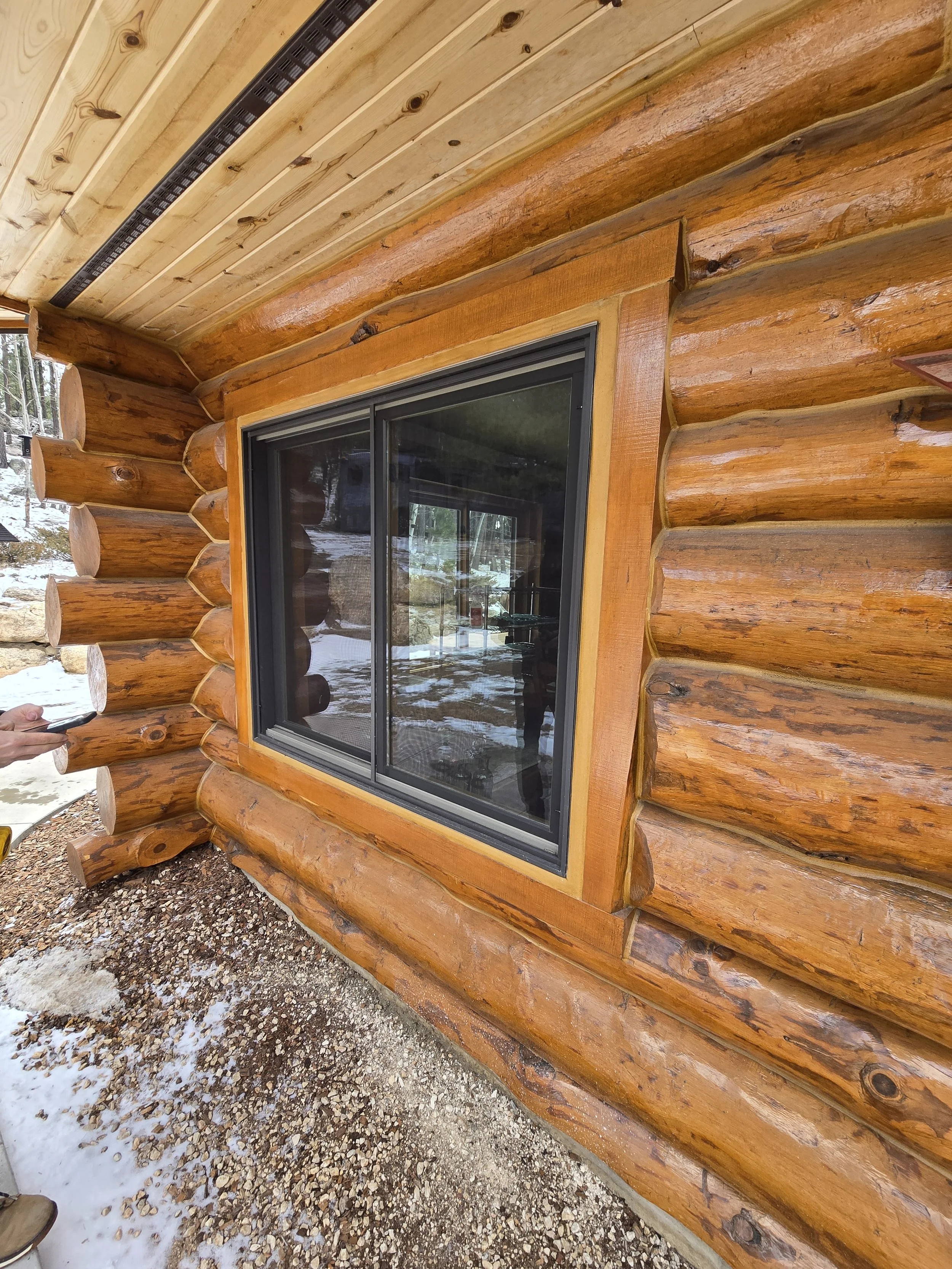 Close-up of a window on a log cabin, showing the logs and the window frame with reflection of trees and a person in a dark shirt.