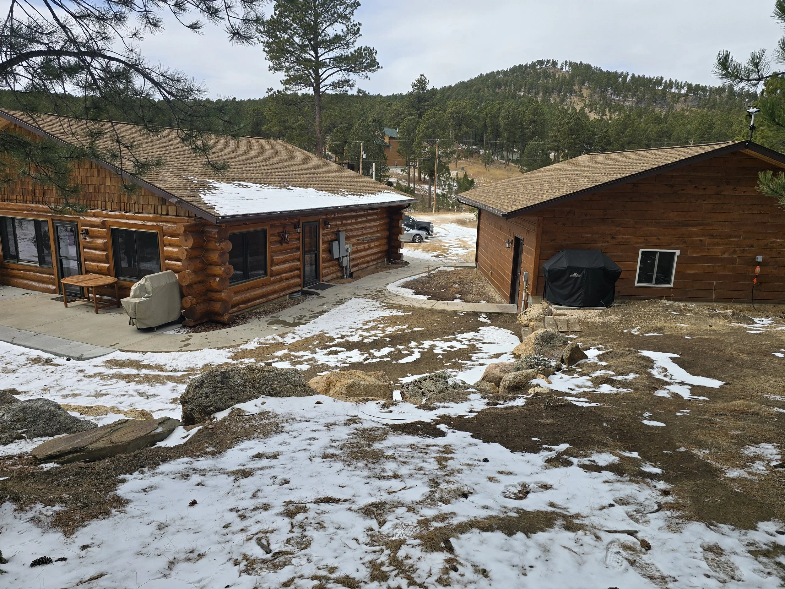 View of two wooden houses in a snowy landscape with trees and hills in the background.