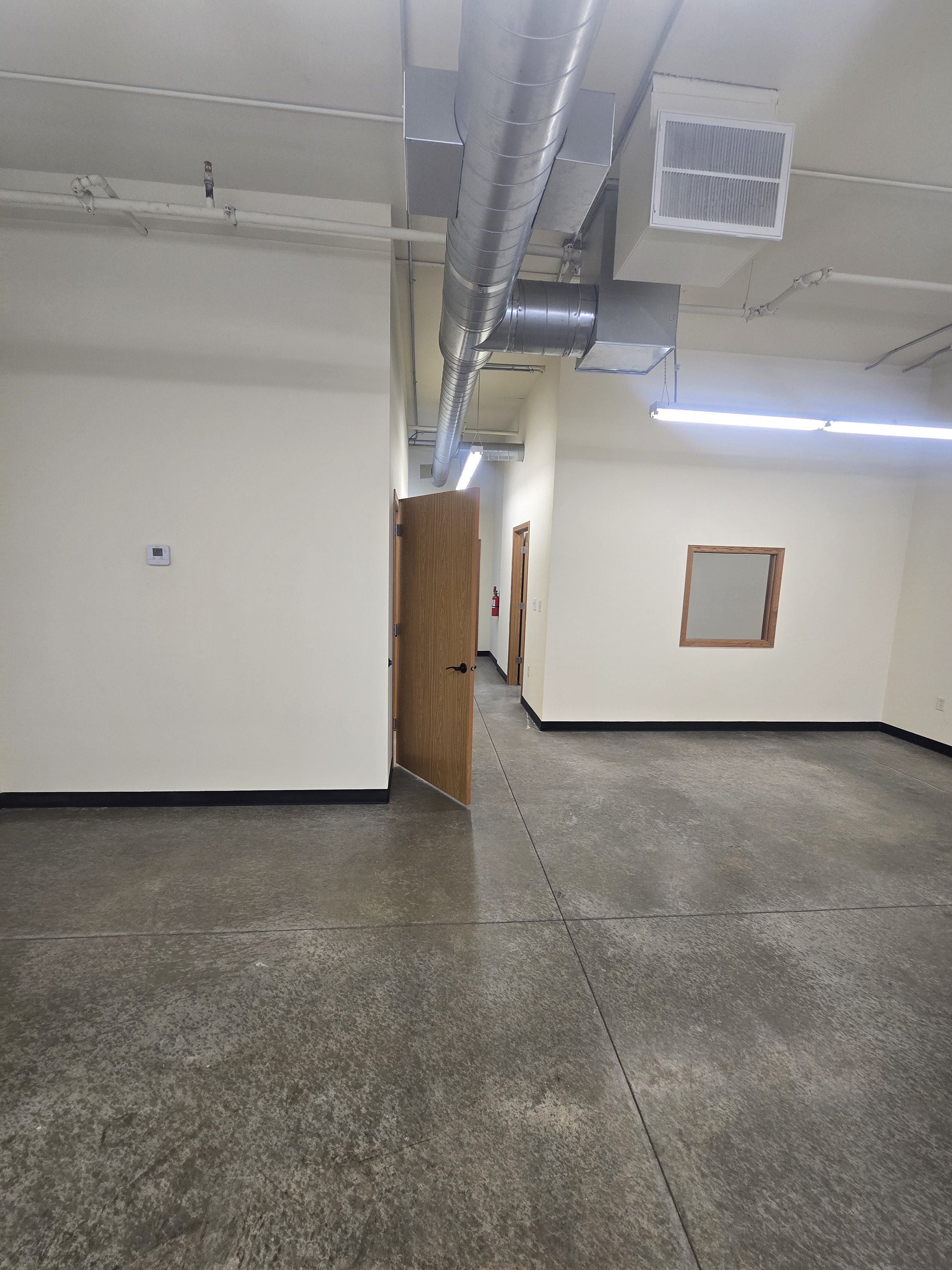 Empty industrial or office space with beige walls, concrete floors, open ceiling with exposed ducts, and partially open wooden door.