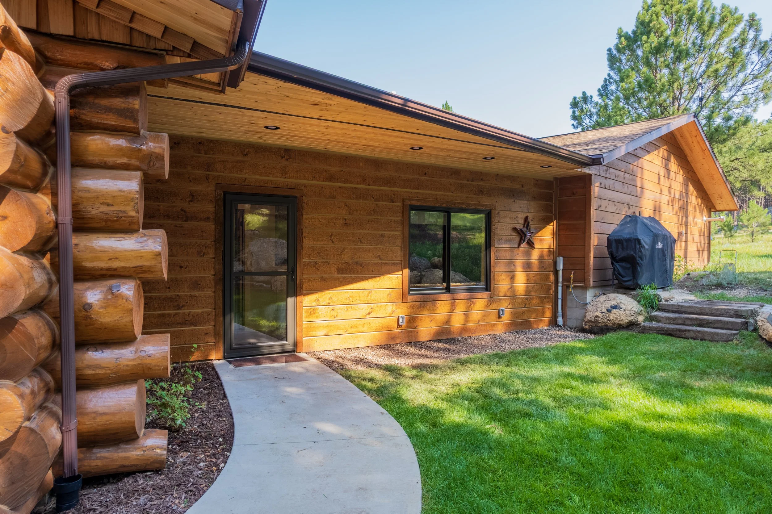 A wooden house with a brown exterior, black-framed glass door, and windows, surrounded by green grass and trees. A concrete pathway leads to the door, and there is a black grill outside. The house has a sloped roof with visible soffit and a star deco