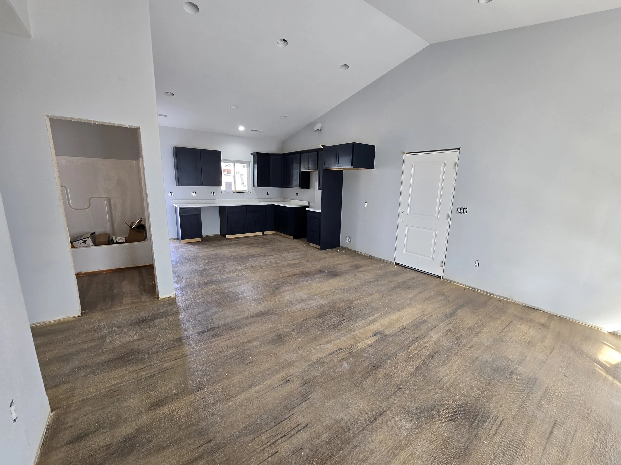 Empty kitchen and living area in a house under renovation with unfinished wooden floors, white walls, black cabinets, and a white door.