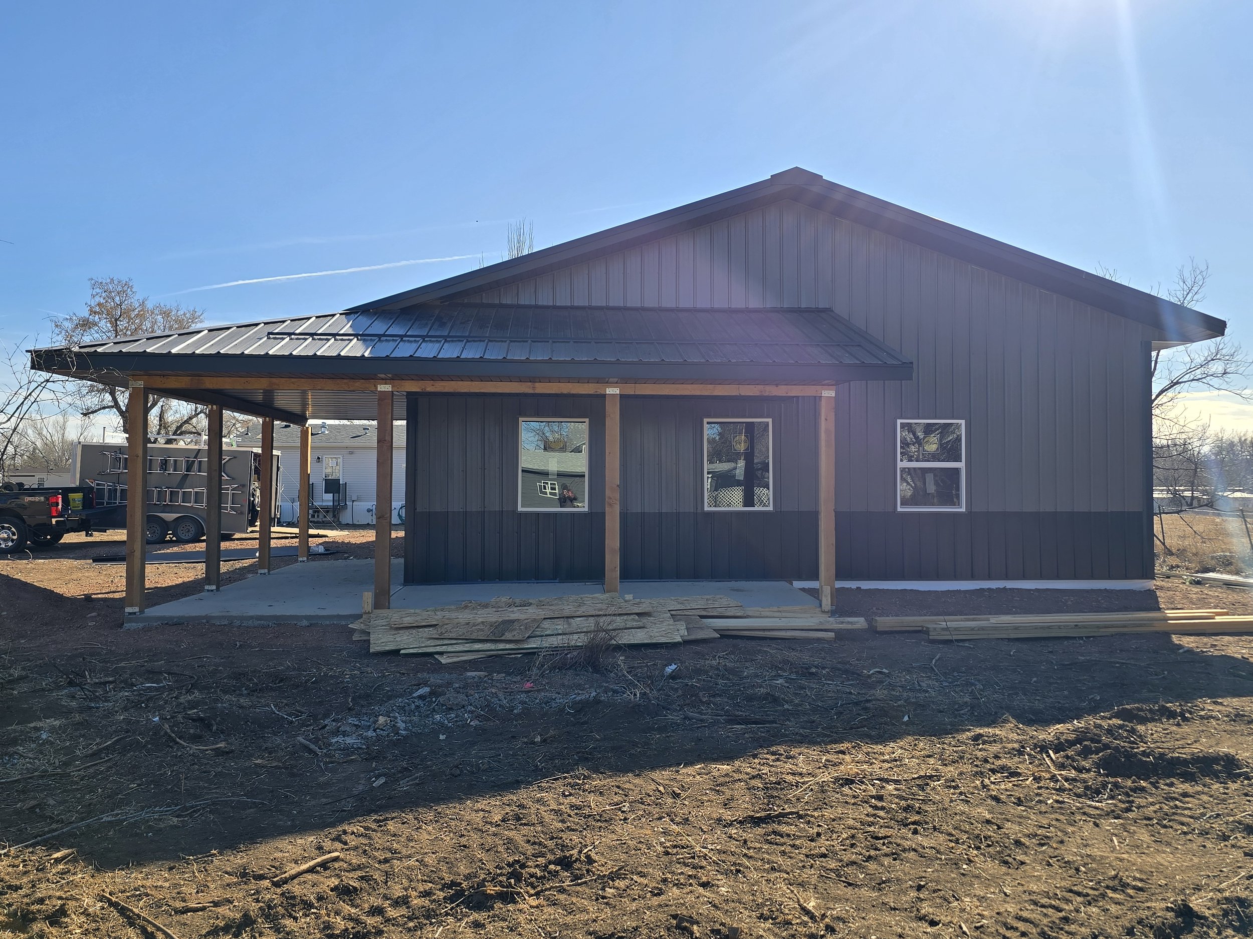Newly constructed house with dark siding, metal roof, and a covered porch under construction, with building materials on the ground.