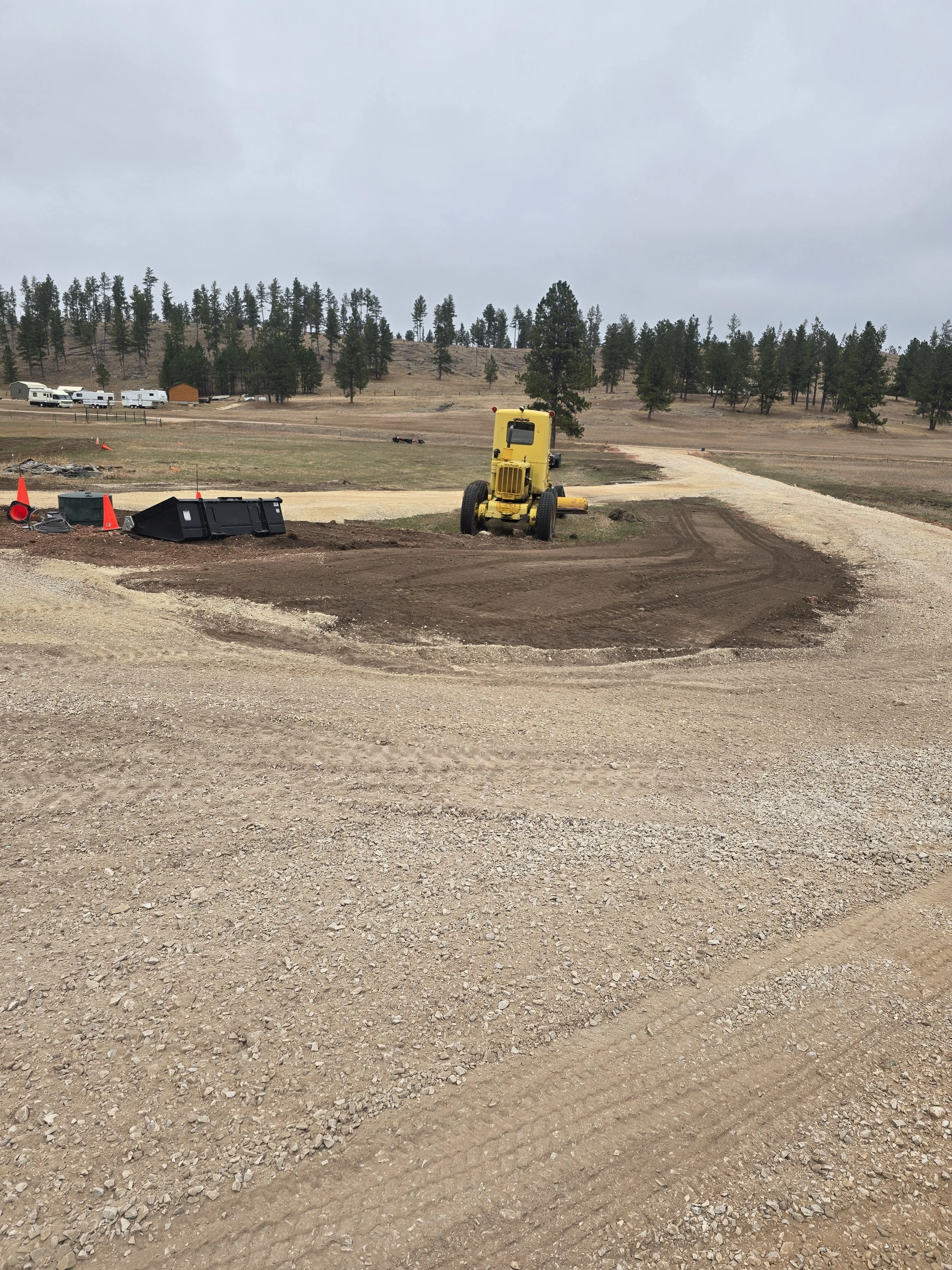 Construction site with a yellow compactor and cleared soil for a new road or driveway in a rural area with trees and parked RVs in the background.