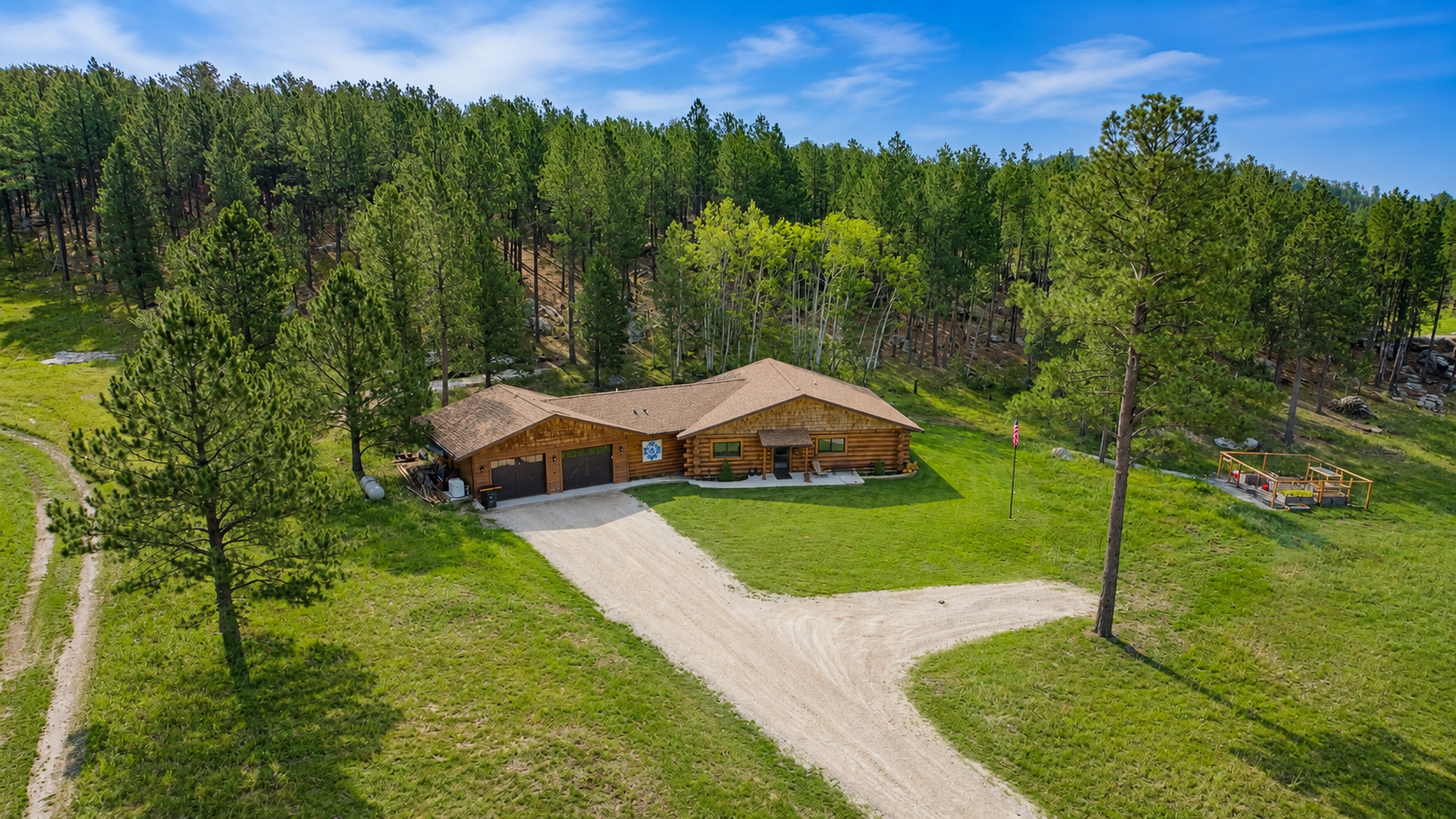 An aerial view of a wooden house in a green, wooded landscape with a dirt driveway leading to the garage, surrounded by trees and open grassy areas.