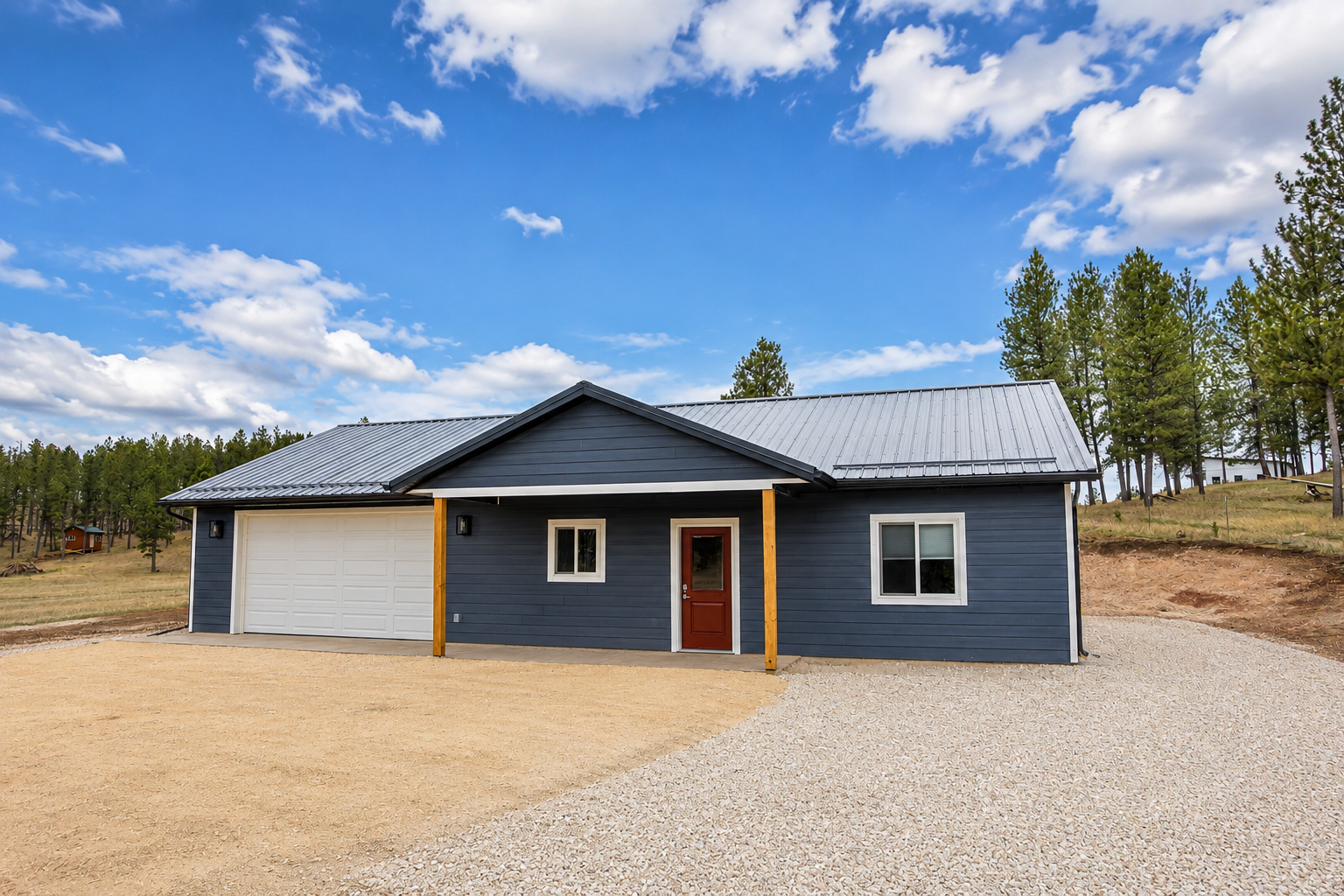 A newly built, blue, single-story house with a metal roof, two windows, and a red front door, set in a rural area with trees and a partly cloudy sky in the background.