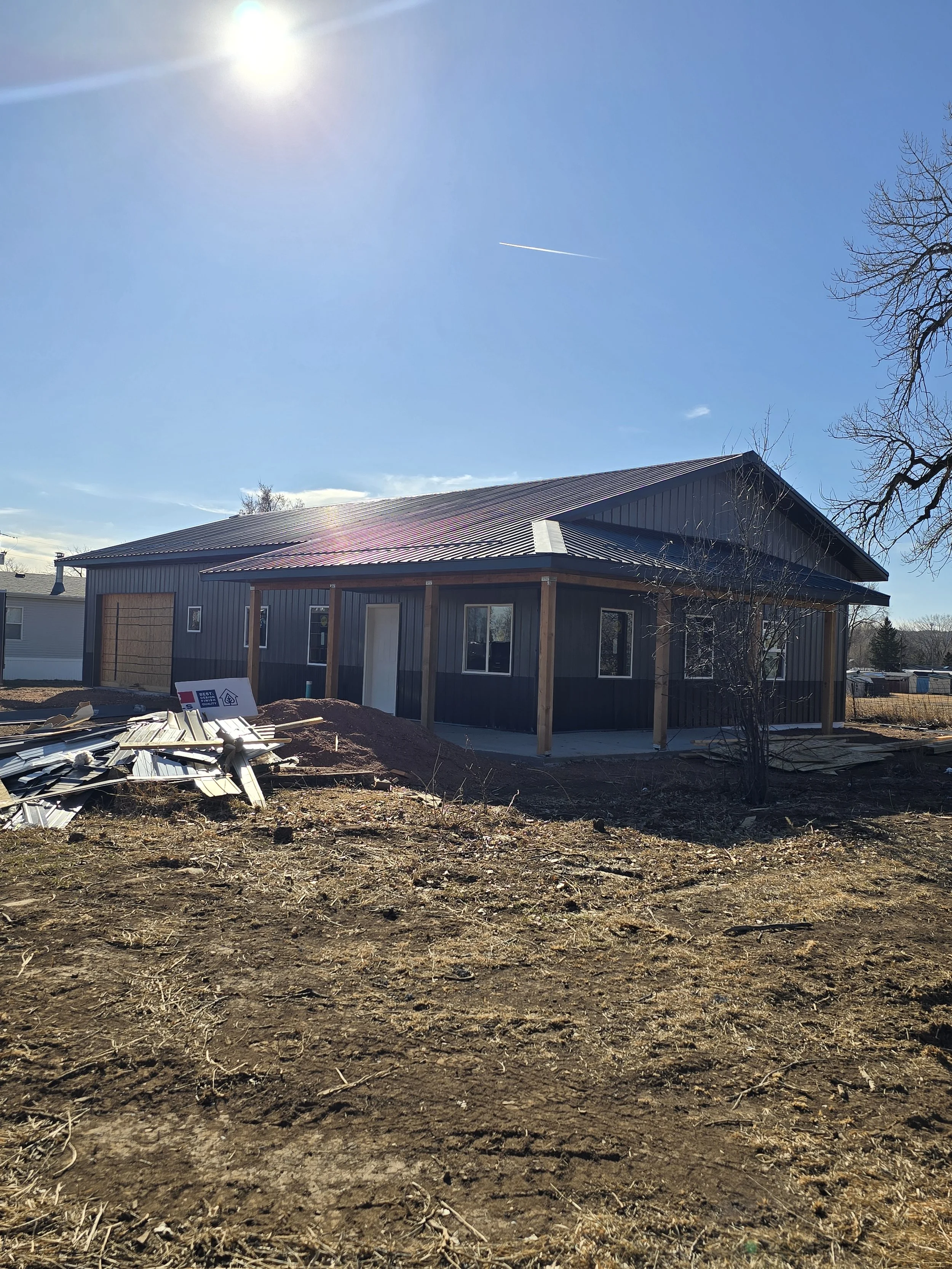 New house under construction with a dark exterior, surrounded by construction materials on the ground, and a clear blue sky with sunlight and a tree on the right side.