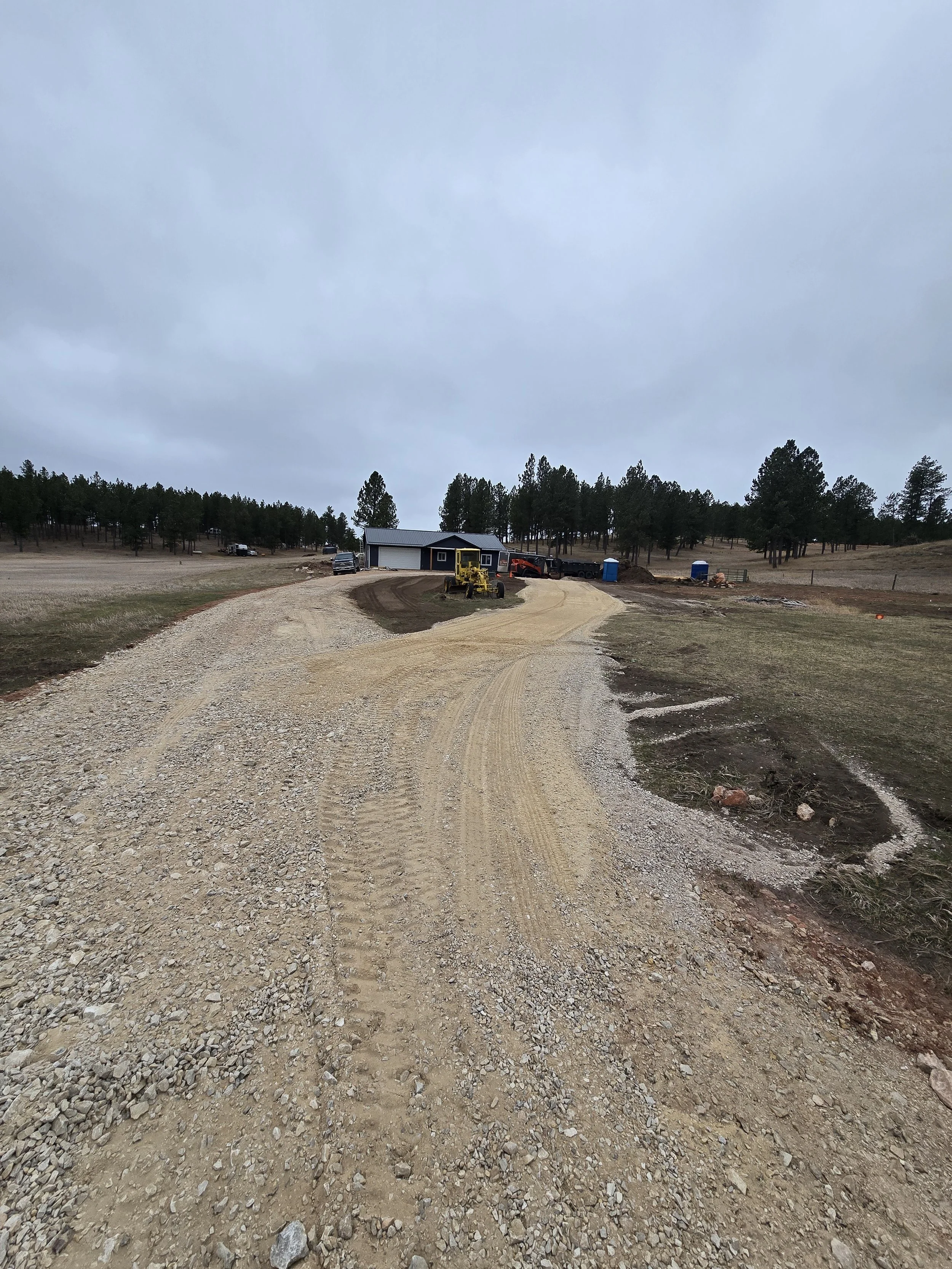 A gravel driveway curves toward a house with a gray roof, surrounded by trees and construction vehicles under an overcast sky.