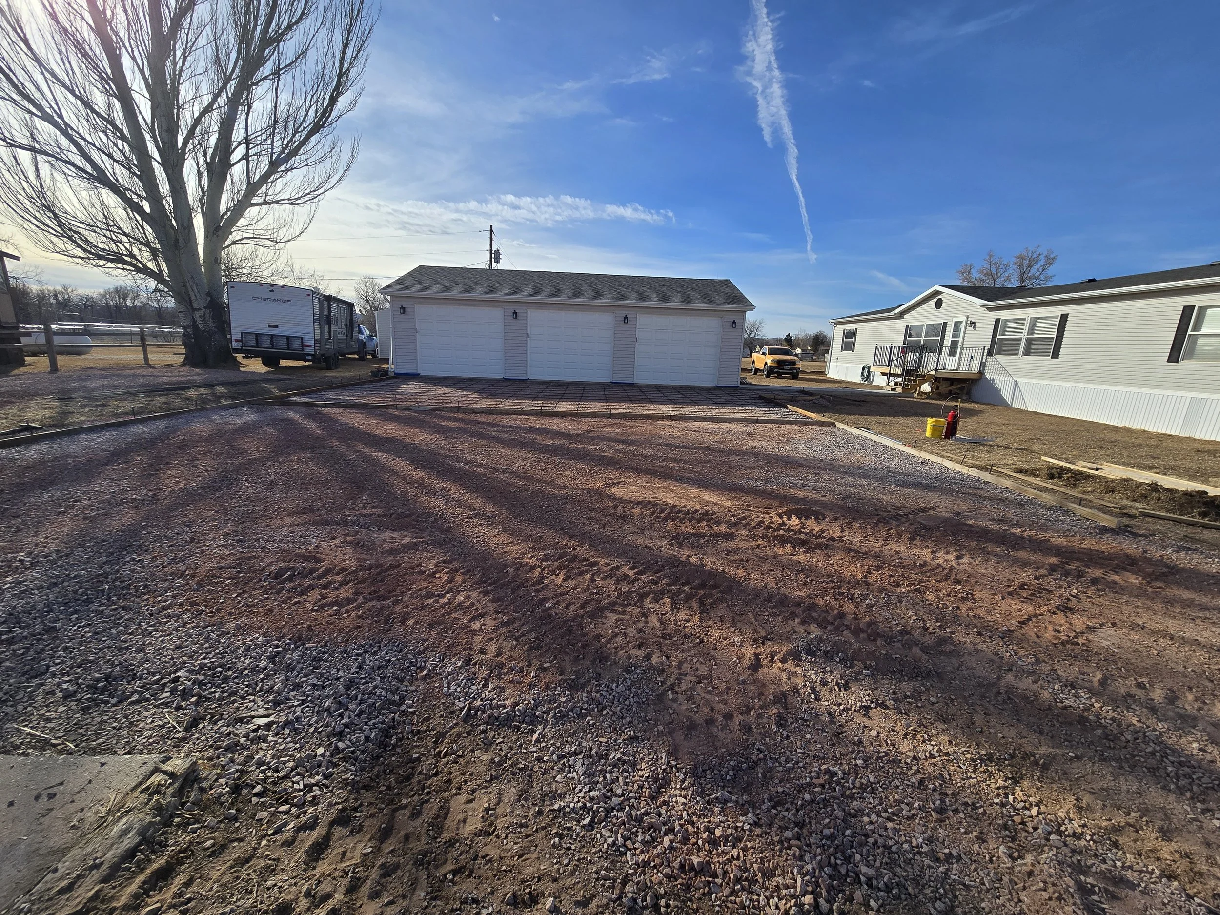 A gravel driveway or yard area being prepared for construction, with a large white garage in the background and a clear blue sky overhead.