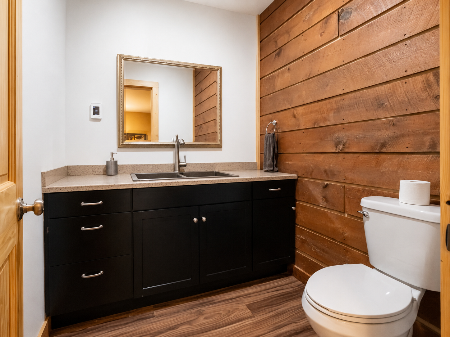 A clean bathroom featuring a black vanity with silver handles, a beige countertop with a double sink, a mirror above, a wall-mounted soap dispenser, a towel ring with a dark towel, a white toilet with a roll of toilet paper on top, and wooden panelin