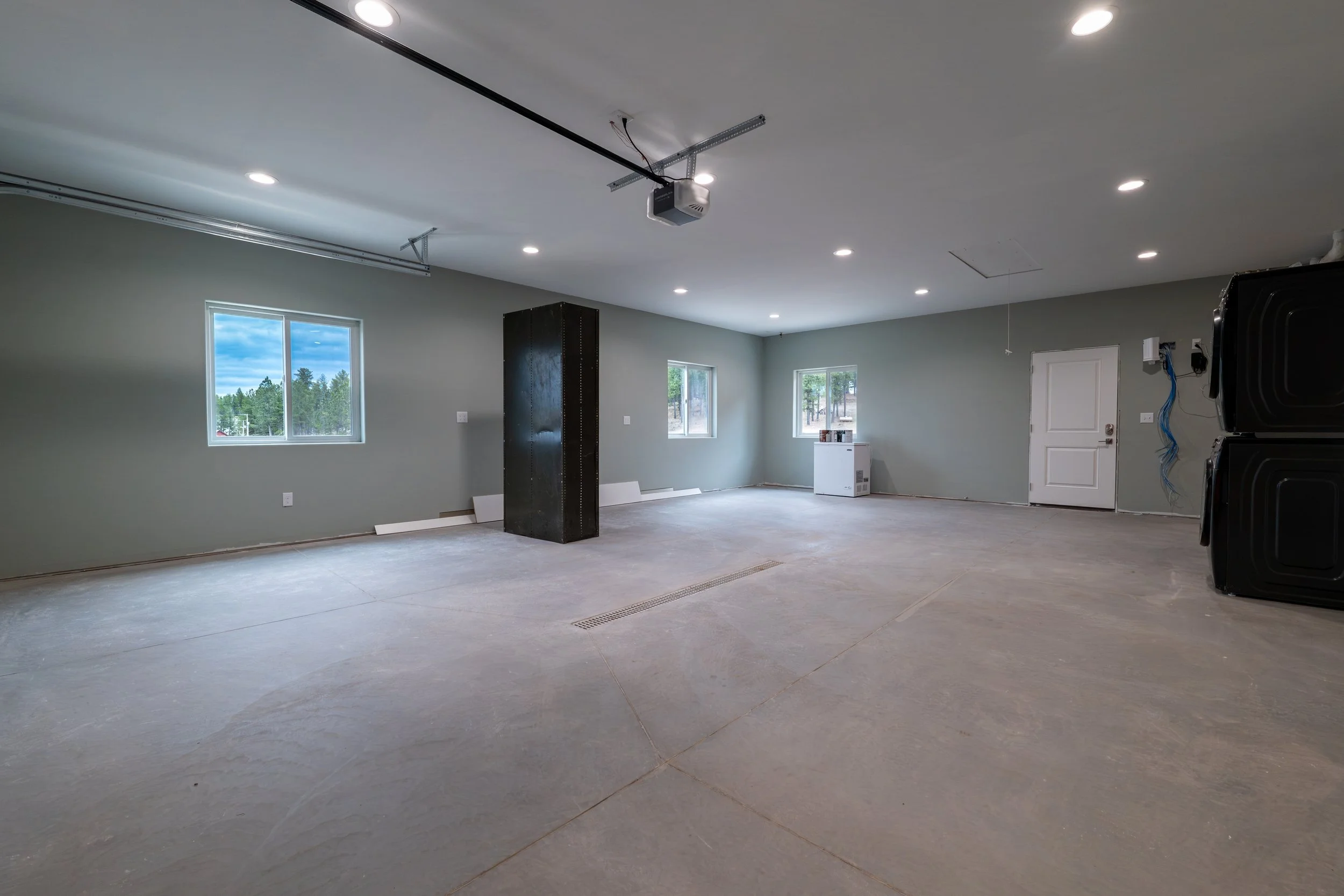 A spacious garage or basement area under construction, with unfinished concrete flooring, three windows letting in daylight, and a garage door opener system installed on the ceiling. The room has gray walls and minimal furnishings.