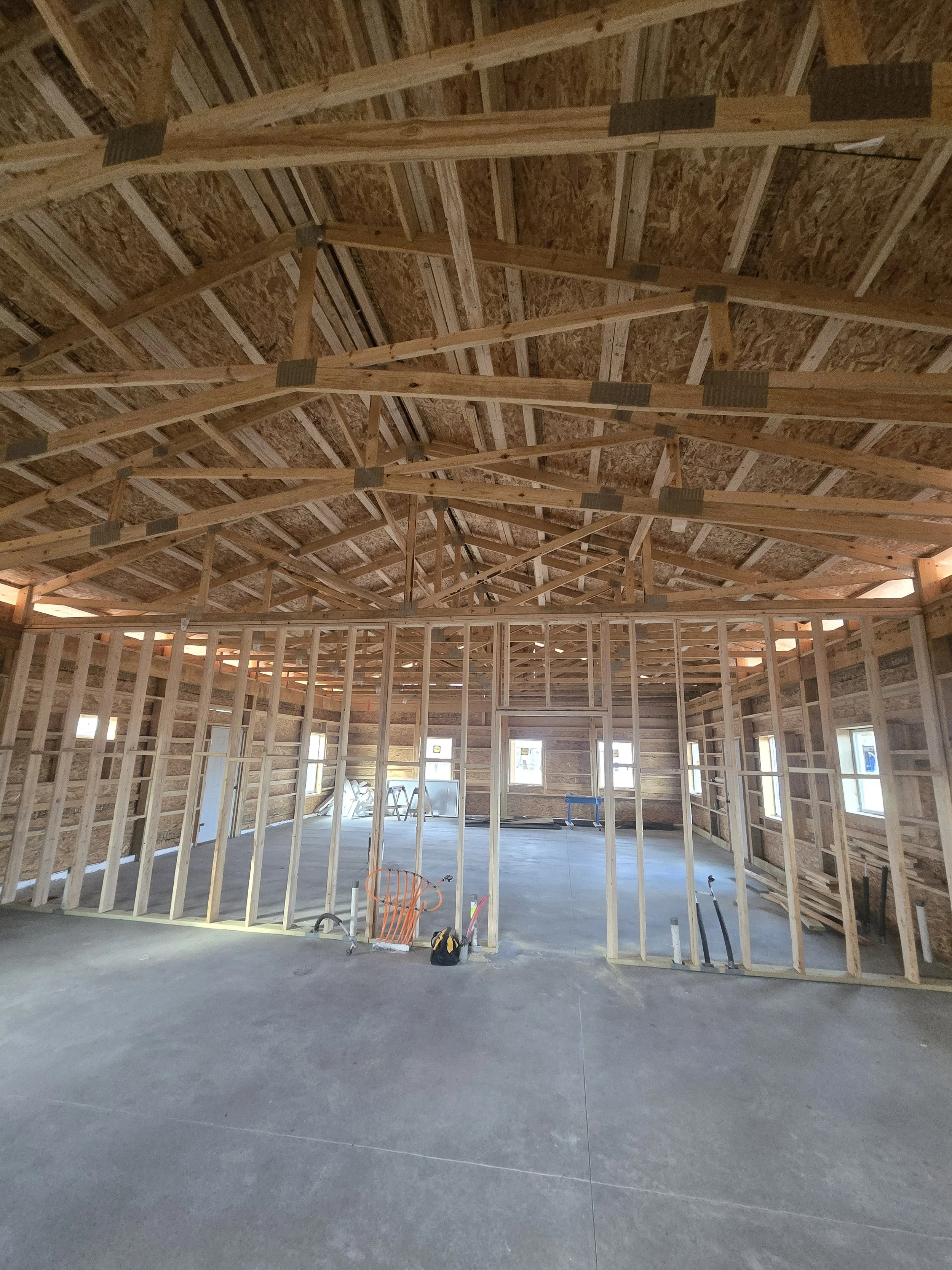 Interior view of a house under construction with exposed wooden framing and a peaked ceiling. Construction tools and materials are on the concrete floor, and windows installed in the background.