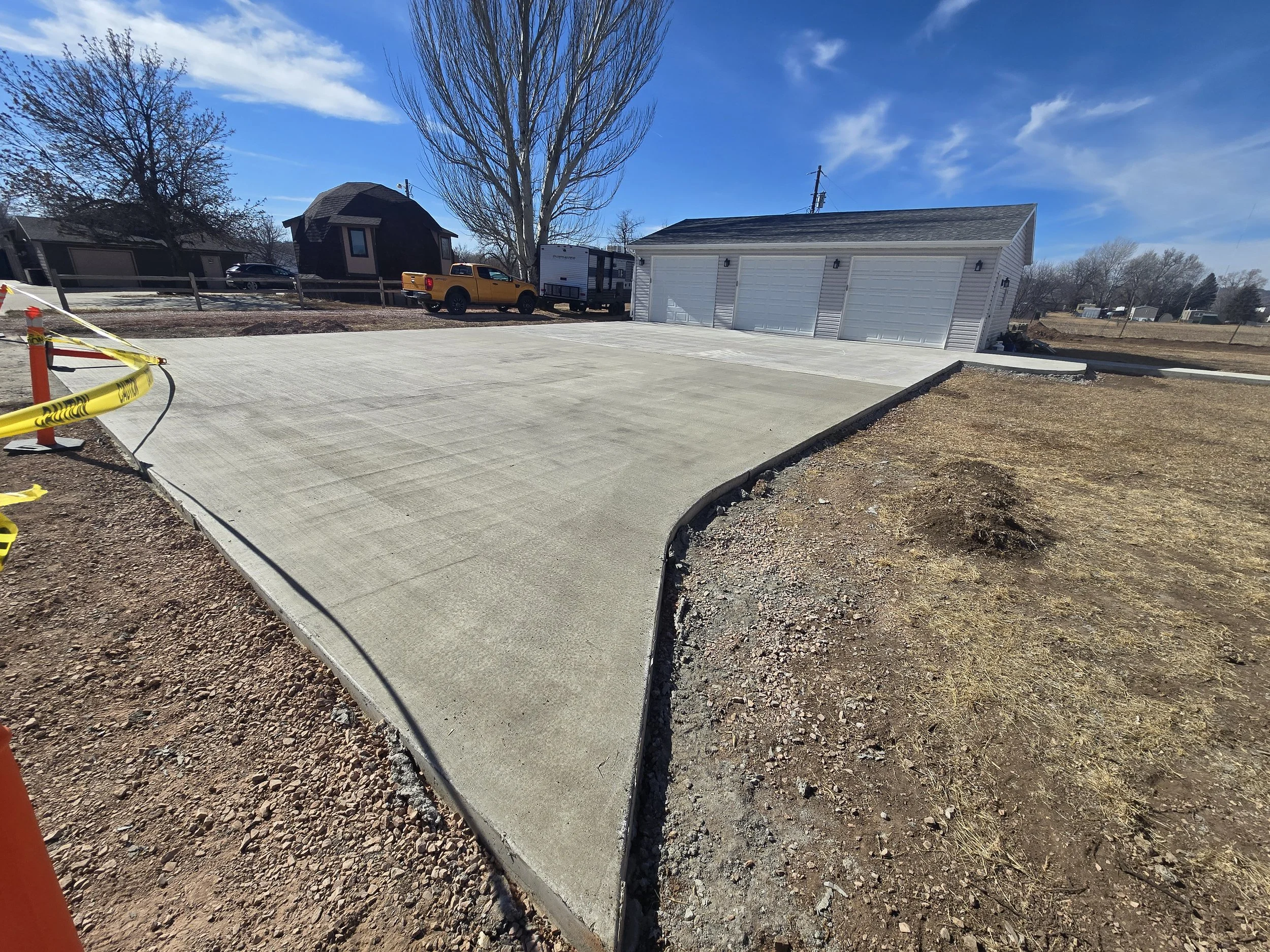Newly poured concrete driveway in front of a garage with multiple white doors, bordered by dirt and grass, with trees and vehicles in the background on a clear, sunny day.