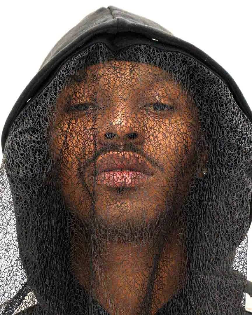 Portrait d'une personne portant une cagoule noire en filet et un hoodie, regardant devant elle.