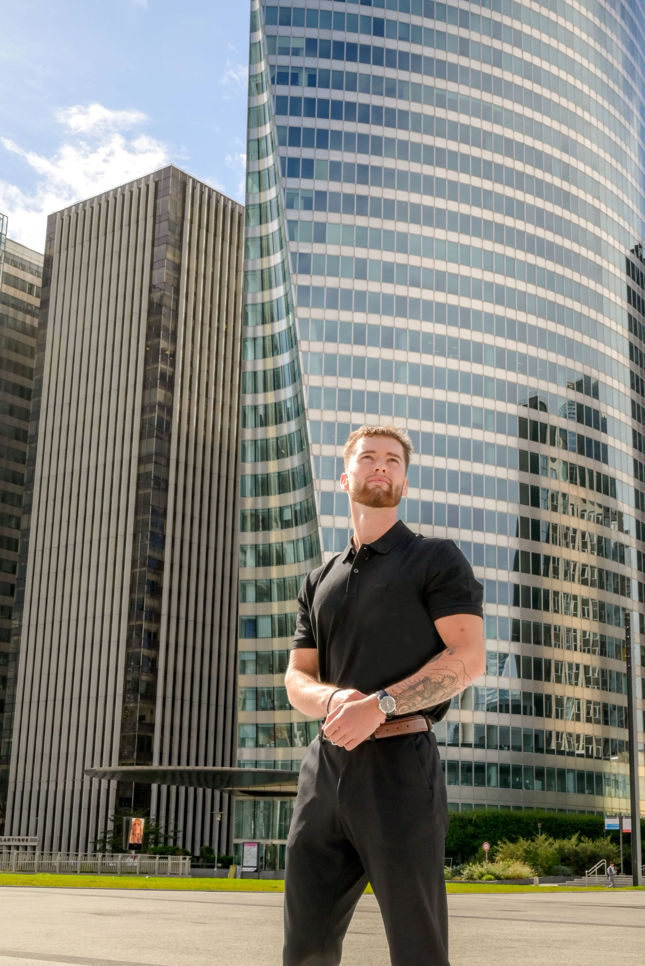 Un homme debout dans un environnement urbain avec de grands bâtiments en verre et une pelouse. Il porte un polo noir, une montre, a une barbe et des tatouages au bras. Le ciel est dégagé avec quelques nuages.