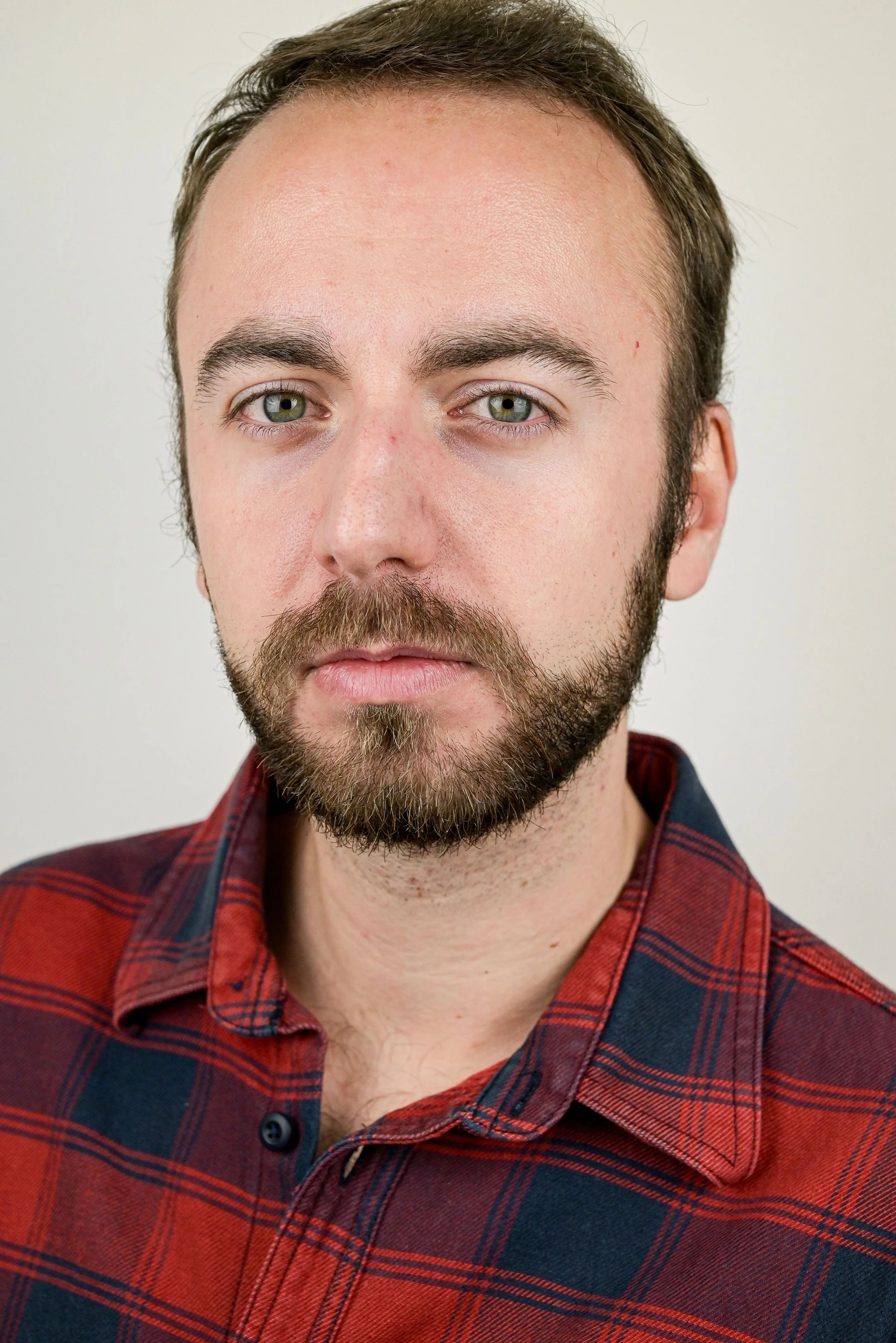 Portrait d'un acteur avec barbe, portant une chemise à carreaux rouges et bleus, fond neutre.