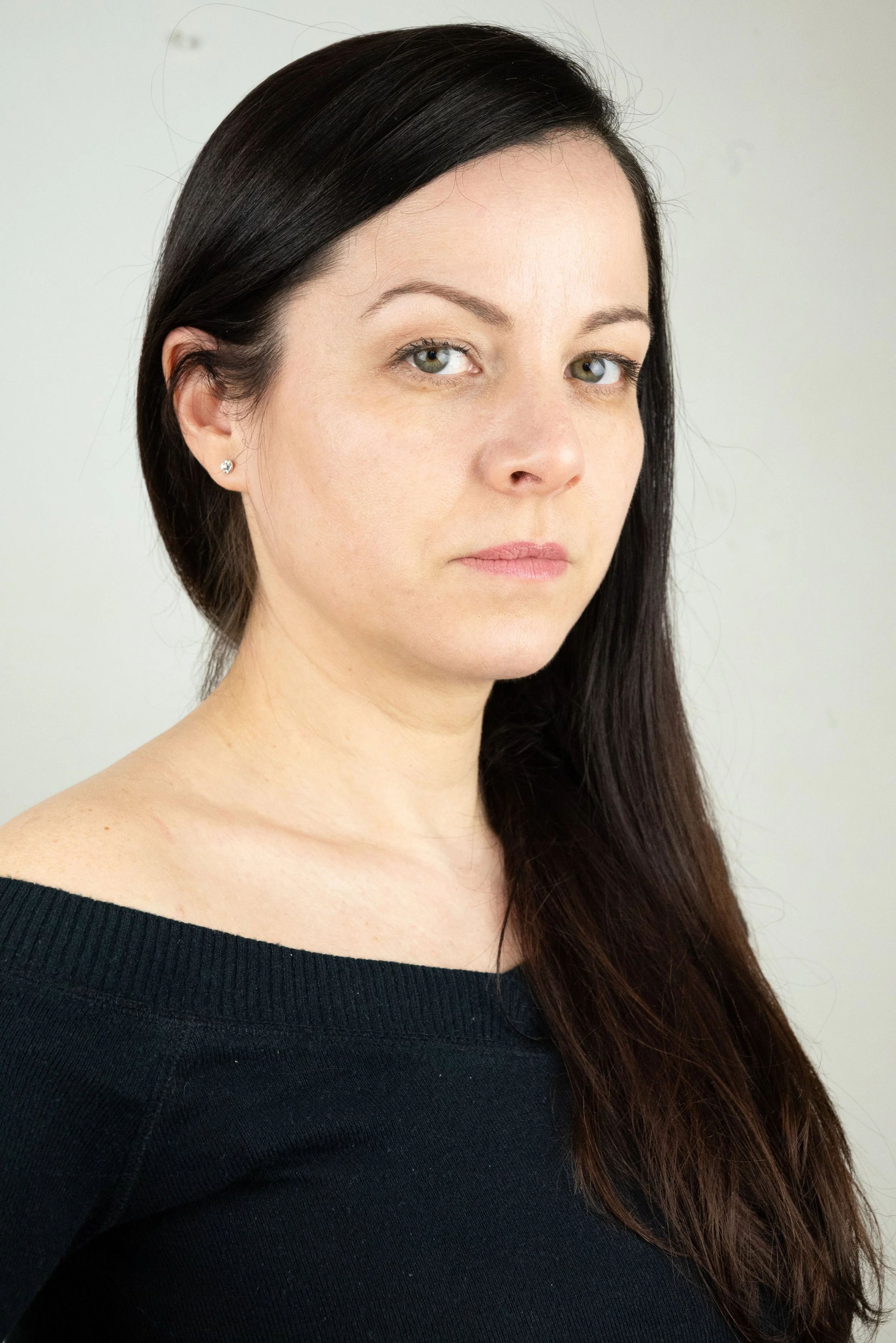 Une actrice aux cheveux longs et noirs portant un pull noir, regardant vers l'objectif avec une expression neutre, en arrière-plan blanc.