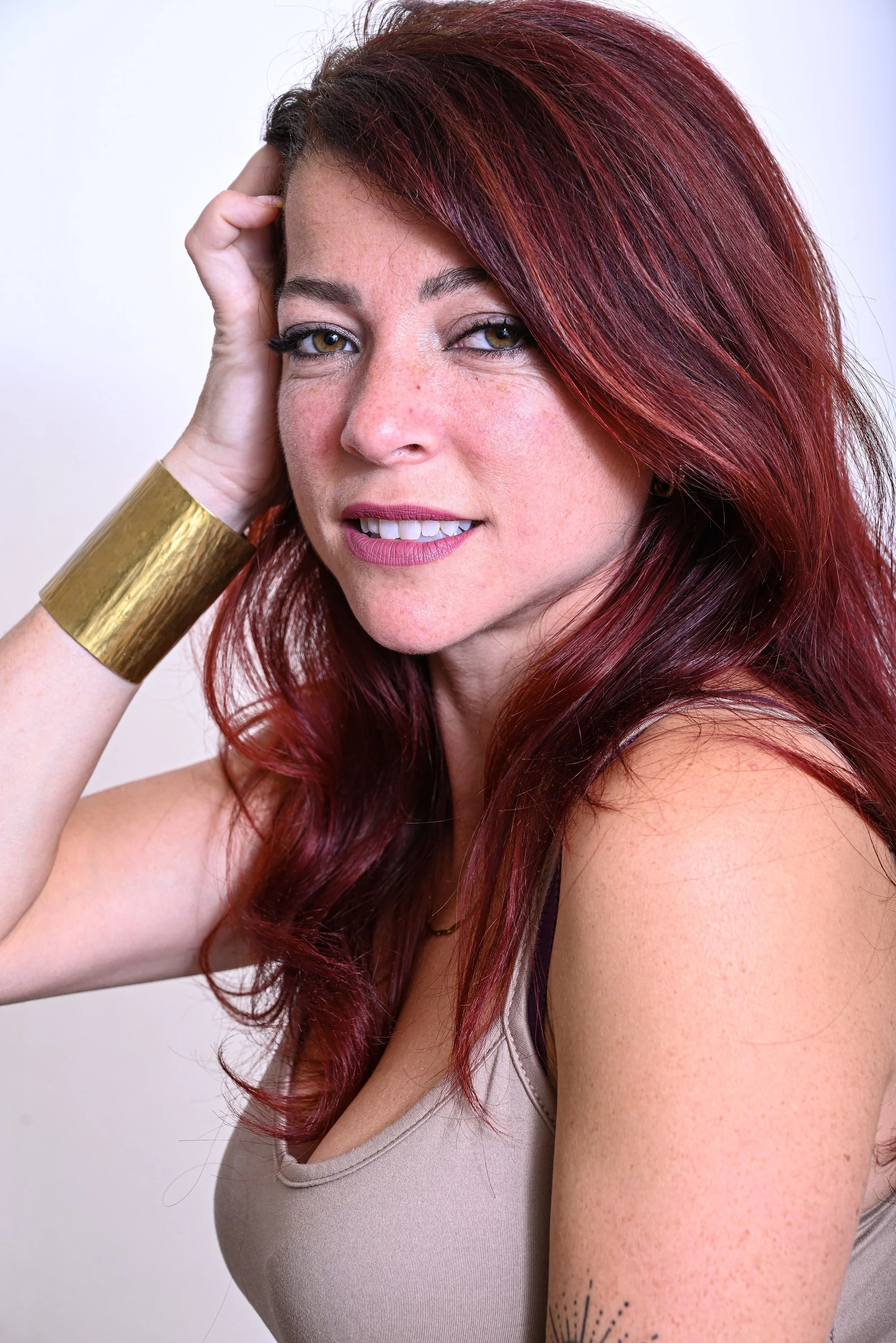 Actrice aux cheveux roux longs et ondulés, portant un bracelet doré, souriant légèrement, en regardant l'objectif, avec un fond blanc.