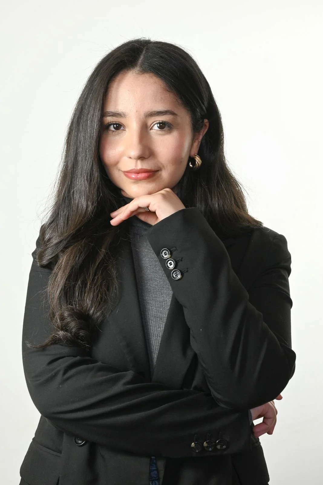 Une jeune femme aux cheveux longs et foncés, portant un blazer noir et un pull gris, pose avec la main sous le menton contre un fond neutre.