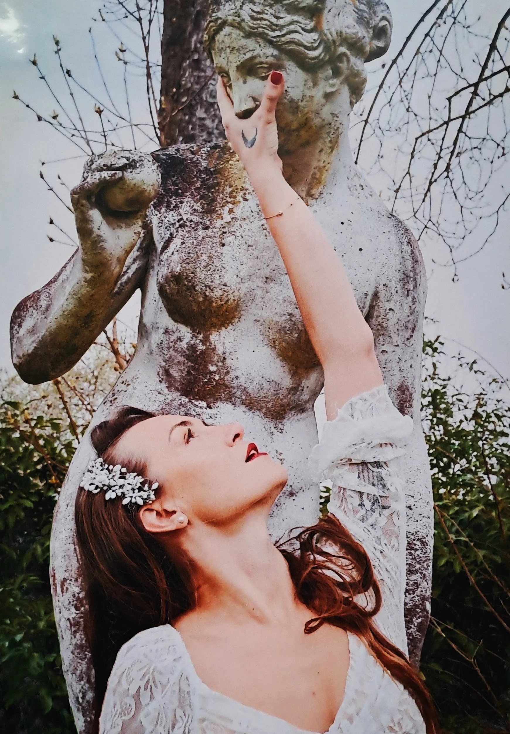 Une jeune femme avec une coiffure ornée de fleurs regarde vers un vieil arbre en posant une main sur une sculpture de statue ancienne représentant une femme nue. La femme porte une robe blanche en dentelle, a les cheveux roux et des lèvres rouges.