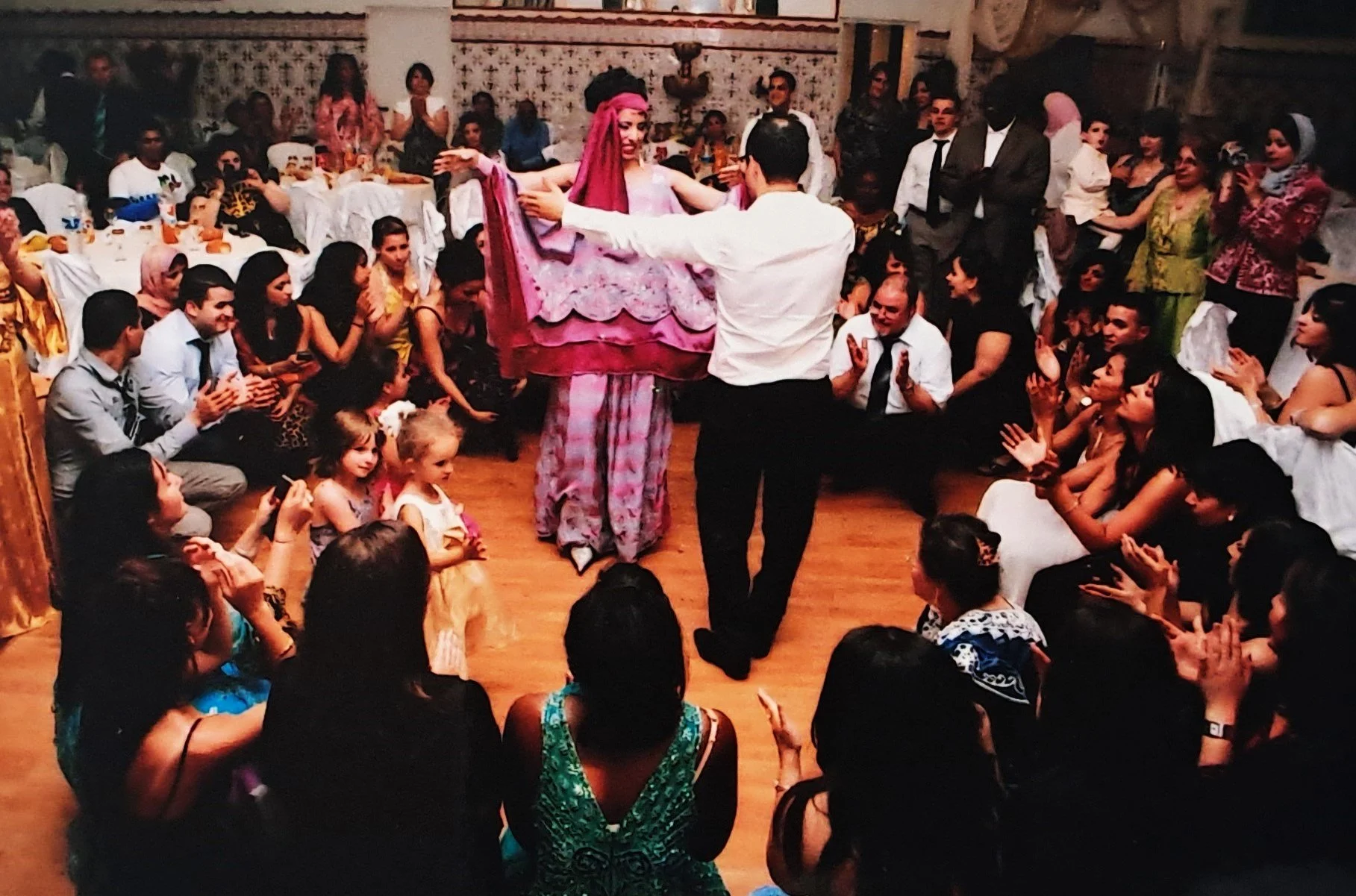 Une célébration ou un mariage avec des invités assis et debout, un homme danse avec une femme sur un tapis, tous portant des vêtements colorés. La scène se déroule dans une salle décorée avec des tables et des sièges.