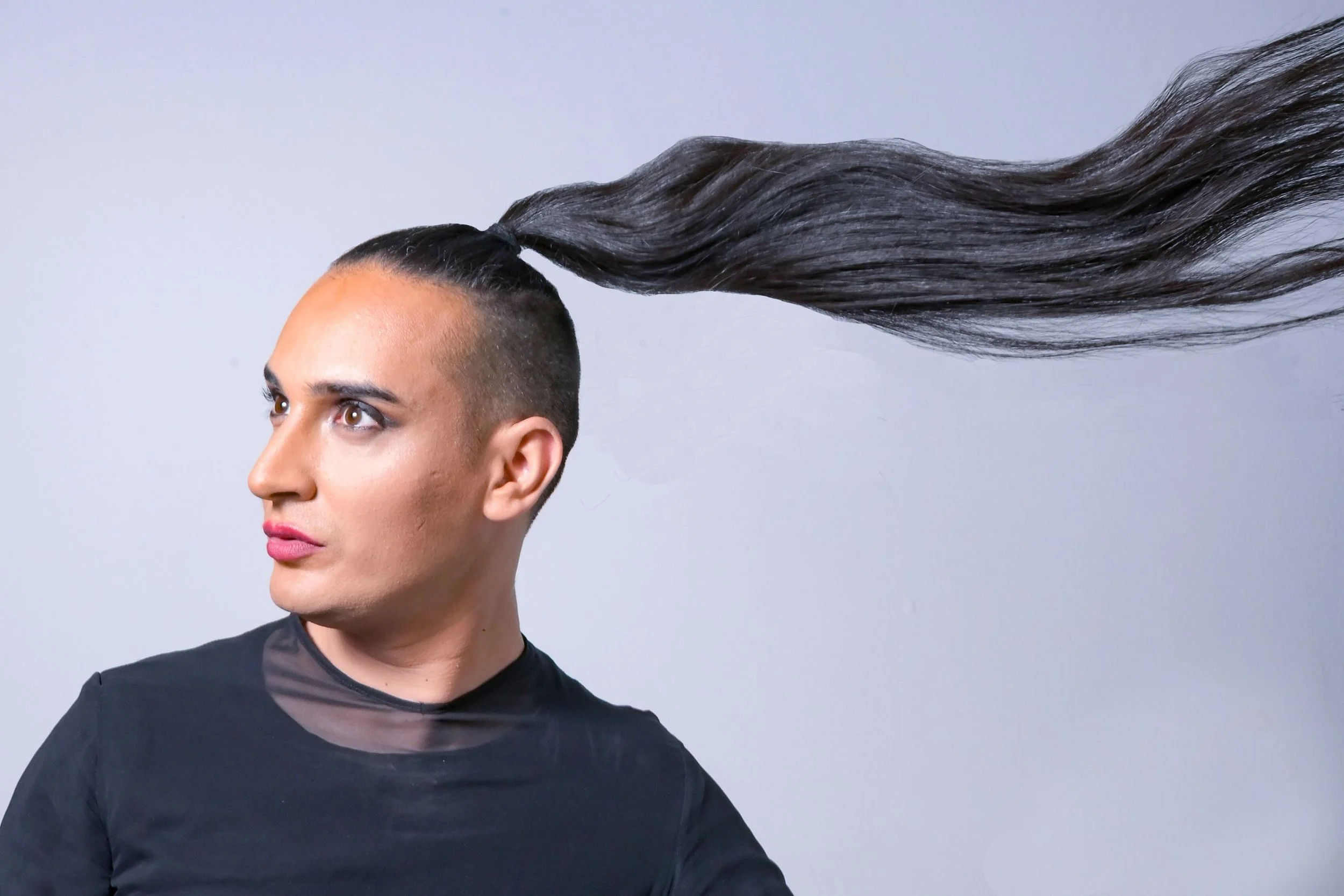 Un artiste avec les cheveux longs attachés, dont la queue de cheval est levée en l'air et s'étend derrière lui, portant un t-shirt noir de face contre un fond gris clair.