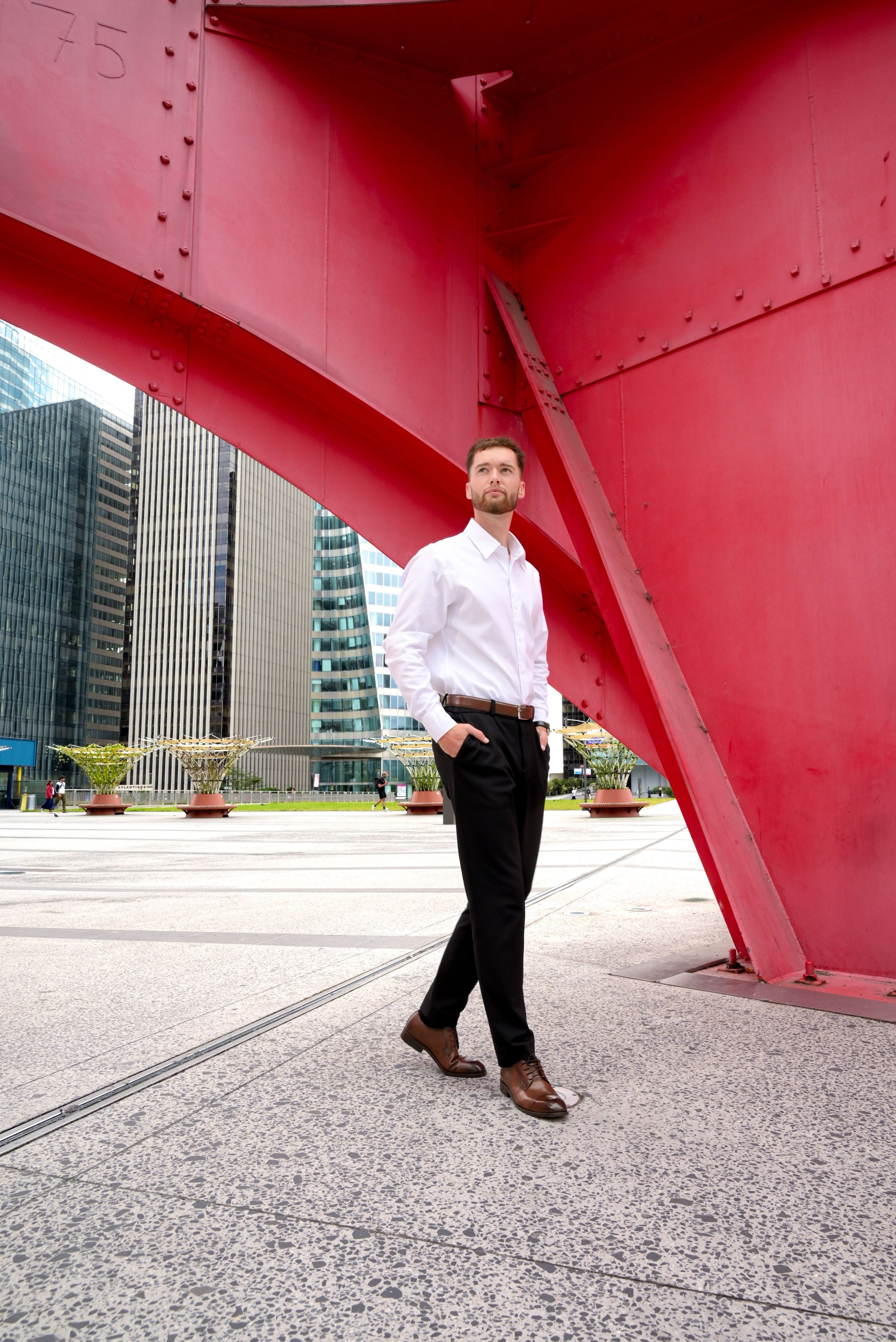 Un homme en chemise blanche et pantalon noir, debout devant une grande structure rouge dans une zone urbaine avec des gratte-ciel modernes en arrière-plan.