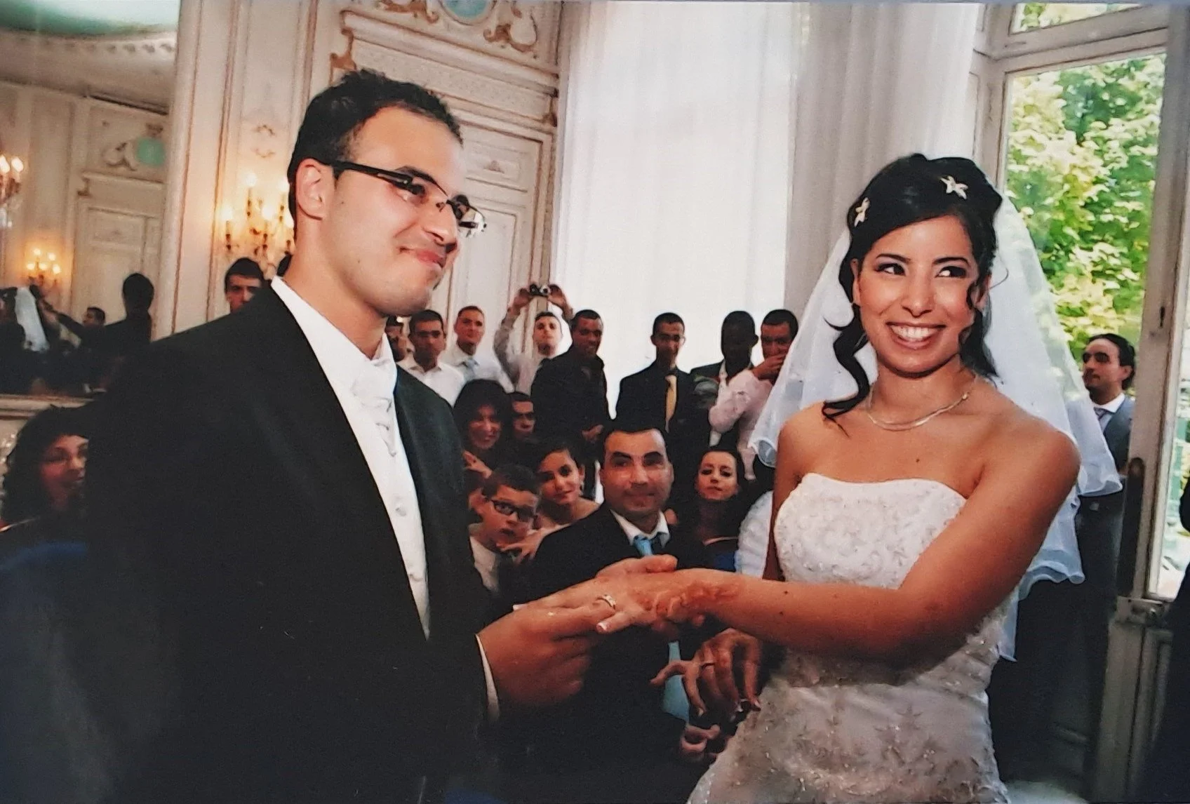 Un couple marié échange des anneaux lors de leur mariage dans une salle ornée, entouré d'invités regardant et prenant des photos.