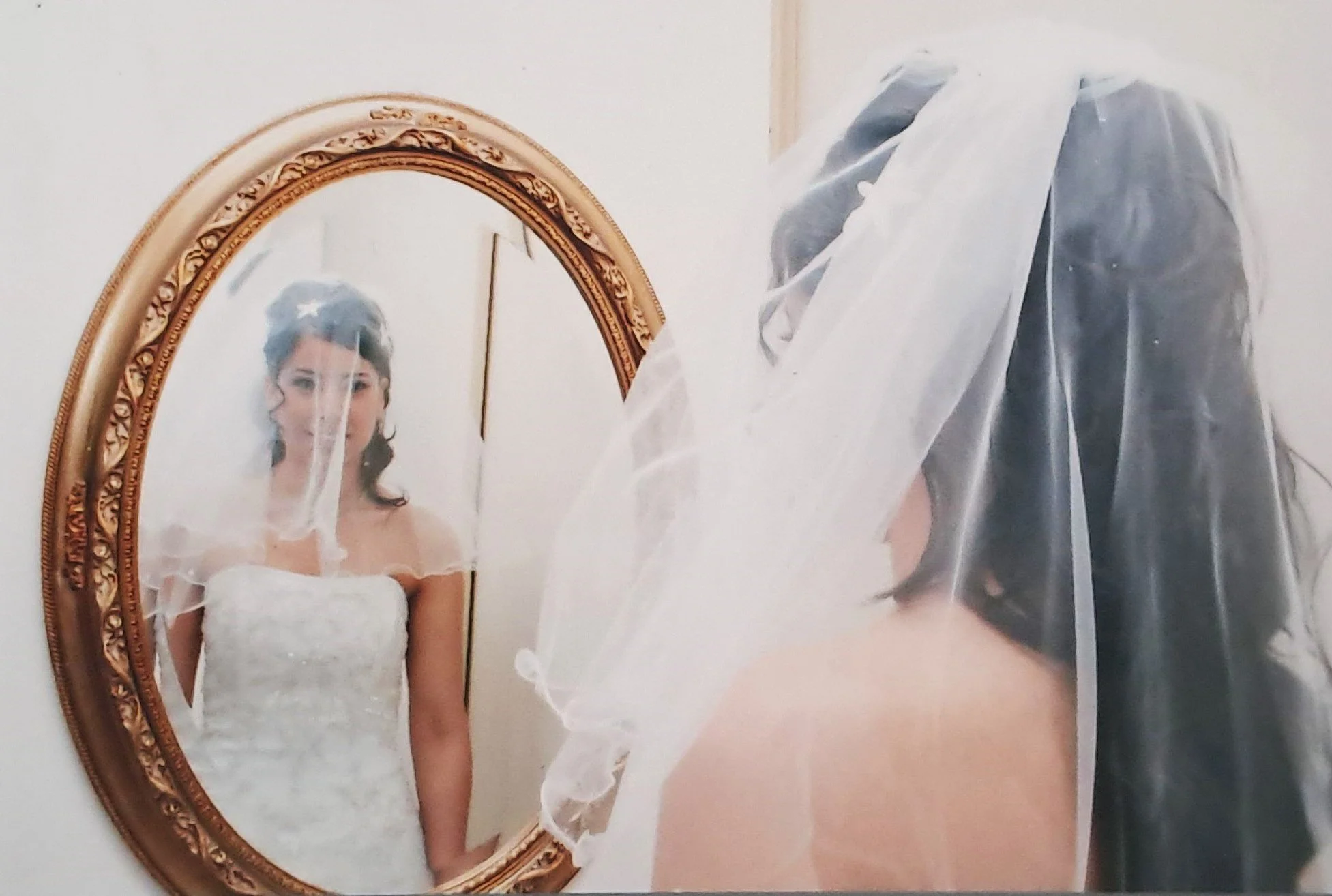 Une femme en robe de mariée regarde son reflet dans un miroir ovale avec cadre doré, portant un voile blanc.