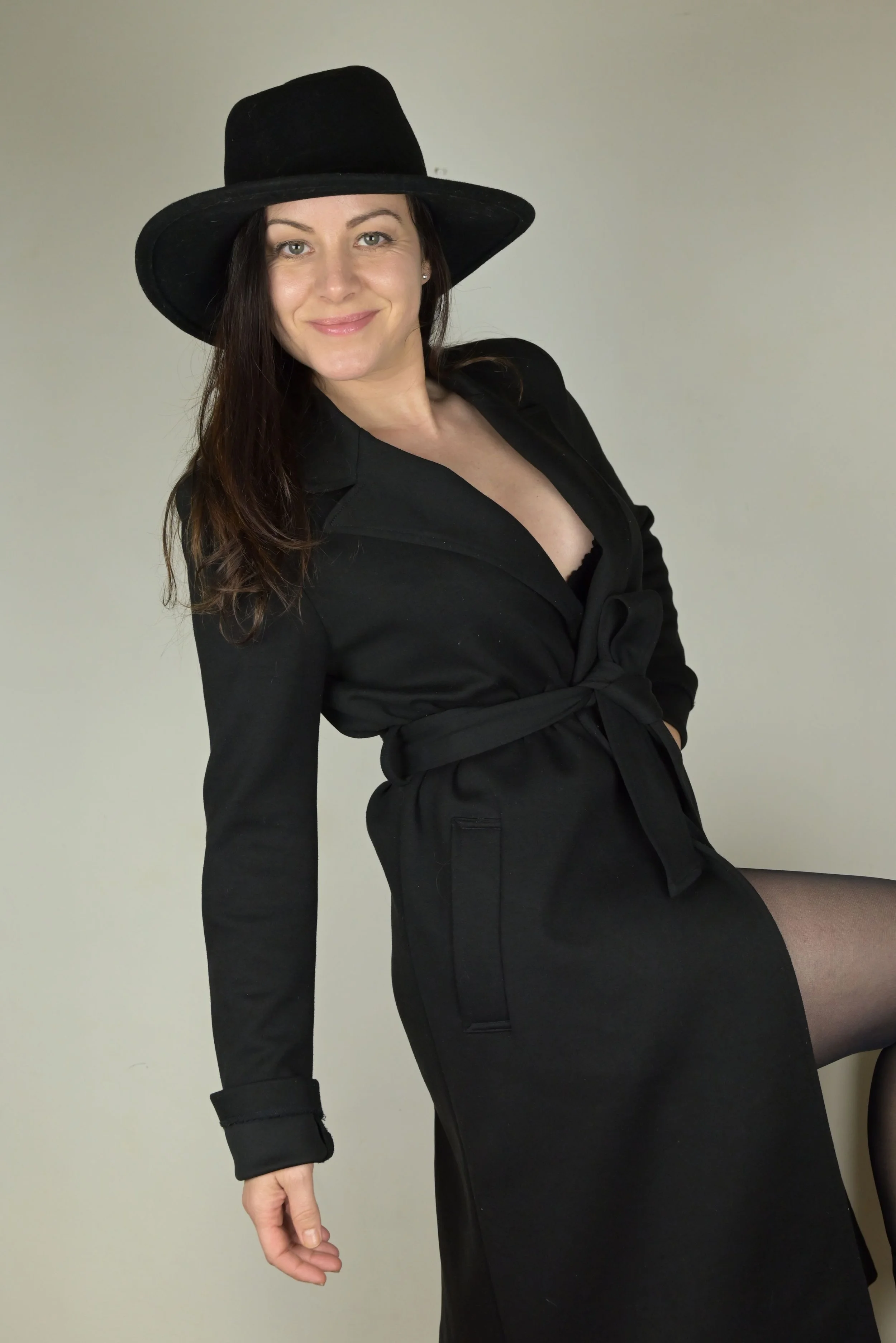 Portrait de l'actrice Sandrine Gauthier, souriante portant un chapeau noir, une robe noire, et des collants noirs, posant contre un mur blanc.