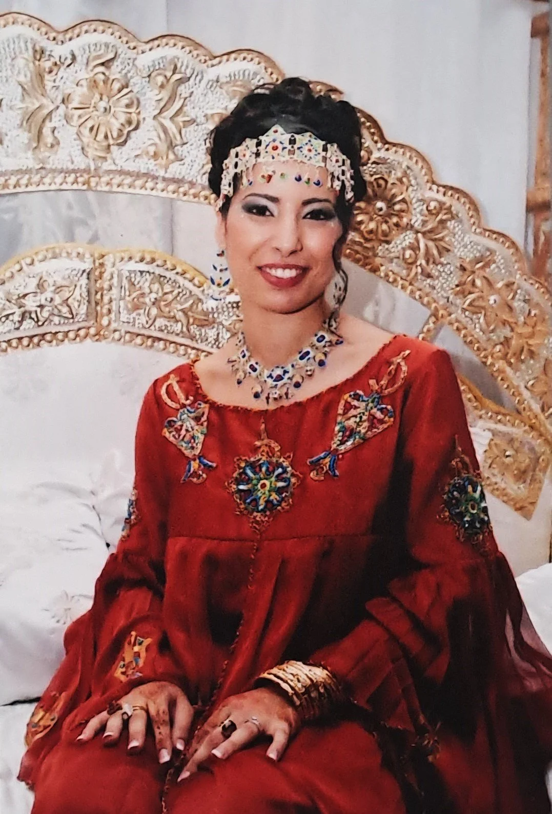 Une femme souriante portant une robe rouge, des bijoux colorés, un diadème et des ornements traditionnels, assise devant un décor doré elaborate.