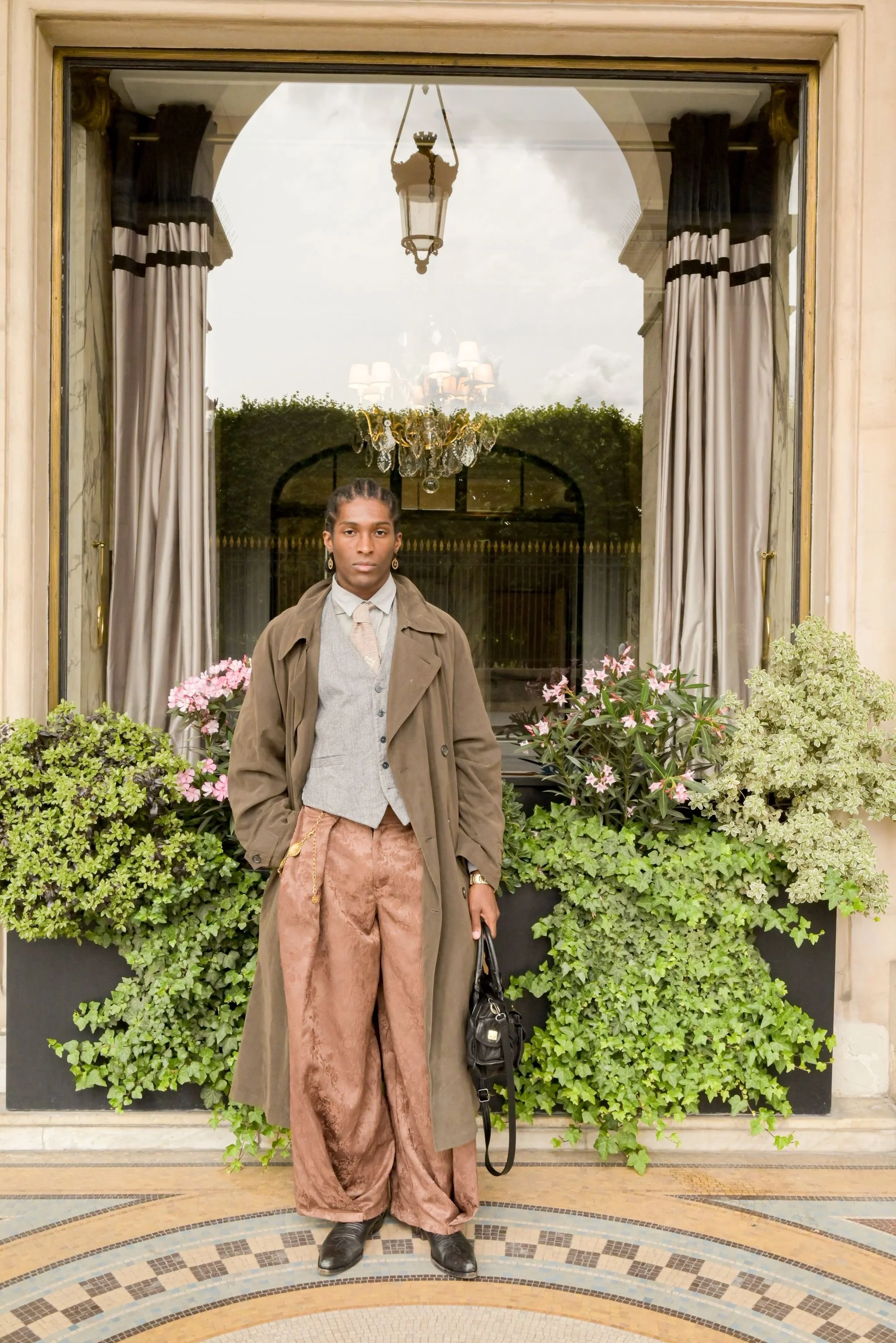 Un mannequin debout devant une grande fenêtre avec des rideaux et une décoration florale, portant une tenue élégante avec un manteau, un gilet, un pantalon en velours et des chaussures en cuir. Séance photo rue de Rivoli