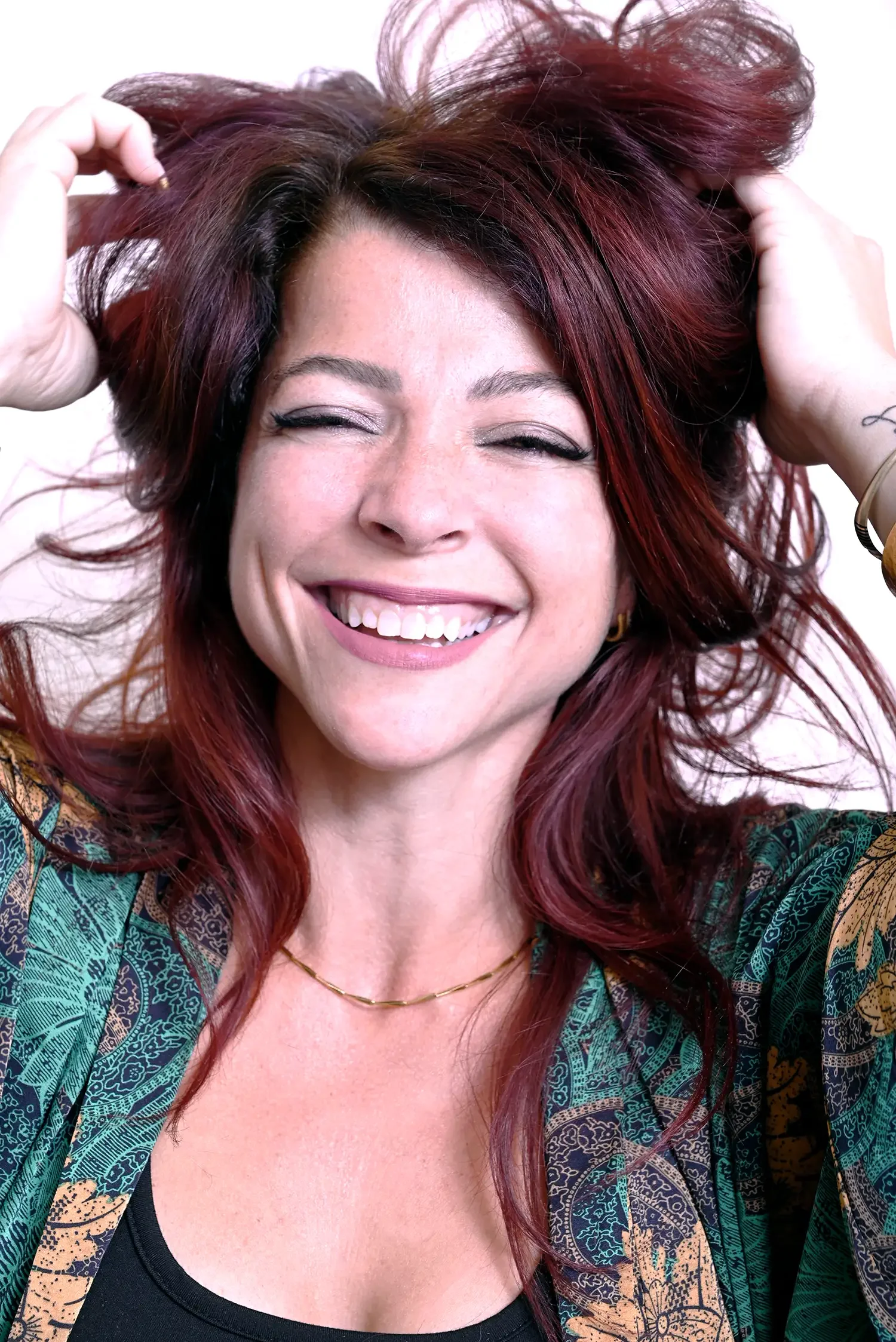 Femme souriante avec cheveux longs et roux, portant un chemisier vert avec motif, souriant largement, fond blanc.