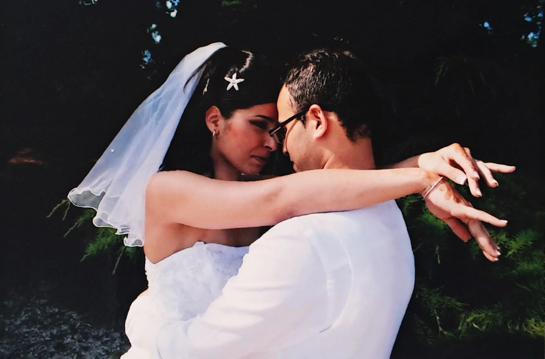 Un couple habillé en blanc, semble lors d'un mariage, se tient de près avec leurs fronts touchant, dans un environnement naturel avec des feuillages verts en arrière-plan.