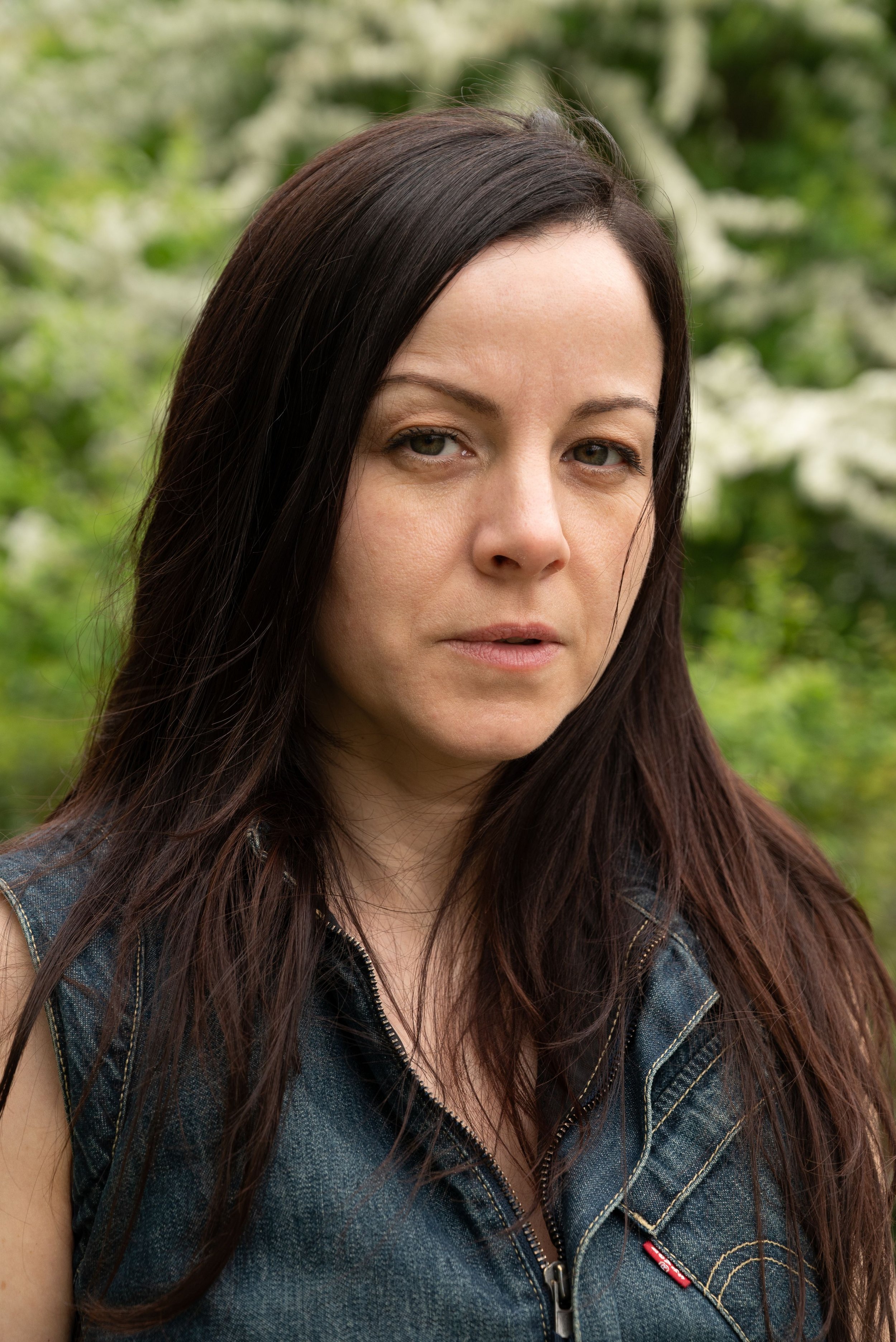 Une actrice aux cheveux longs et foncés, portant un gilet en denim, avec un fond naturel de verdure.