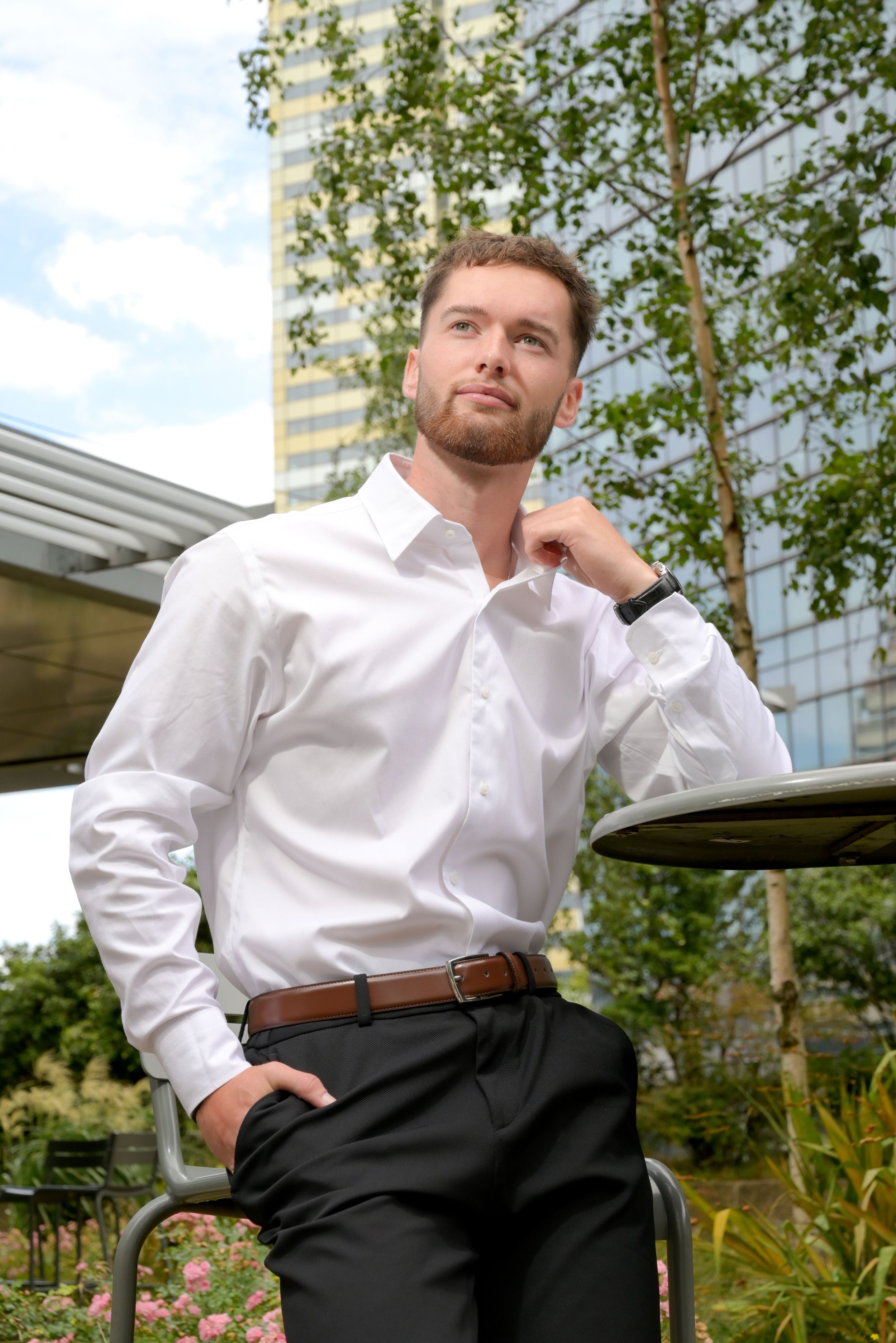 Un jeune homme en chemise blanche, debout à l'extérieur près d'une table ronde, avec un bâtiment moderne en arrière-plan, des arbres et des fleurs.