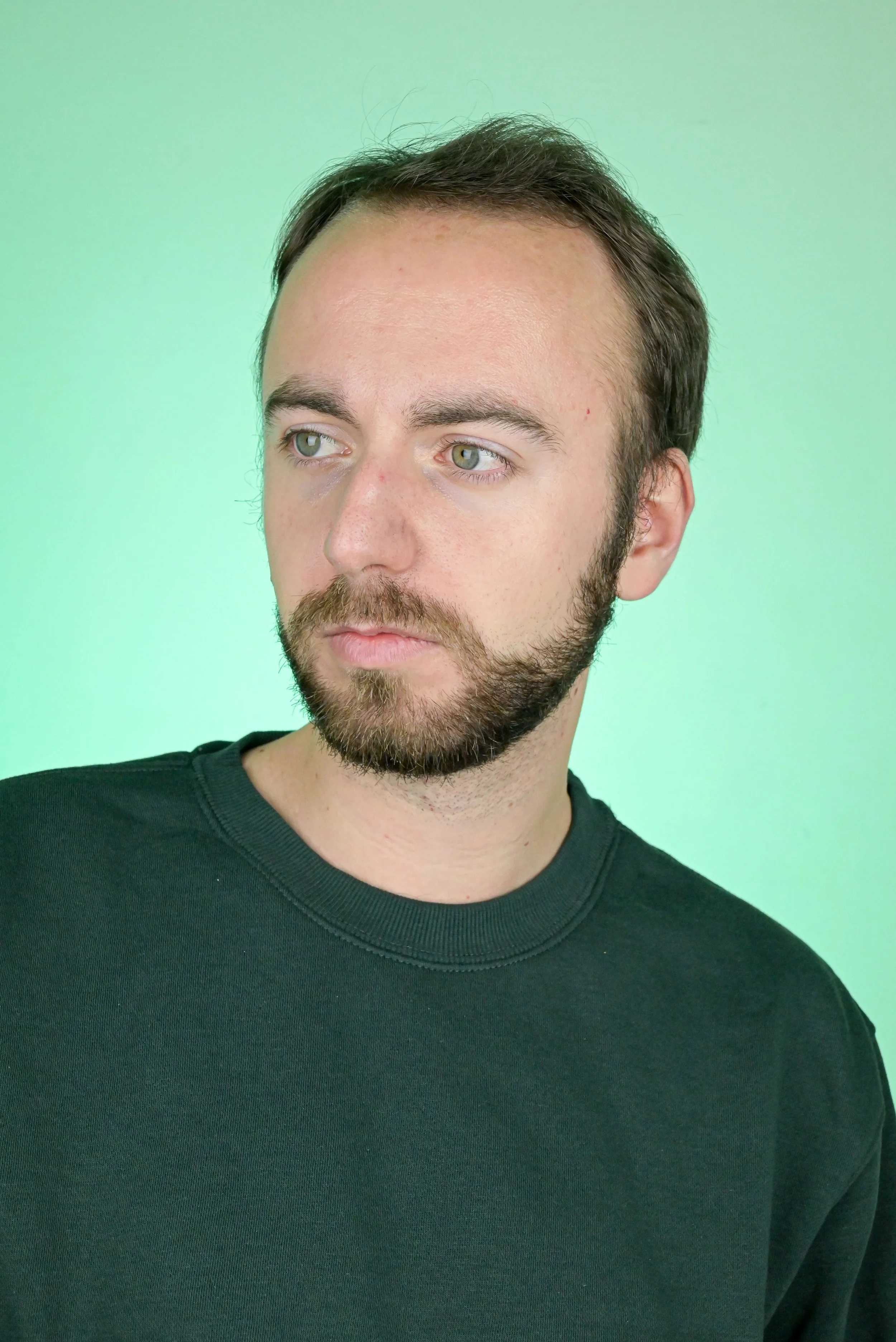 Un acteur avec barbe et cheveux bruns, portant un t-shirt noir, regarde légèrement vers la gauche avec un fond vert clair.