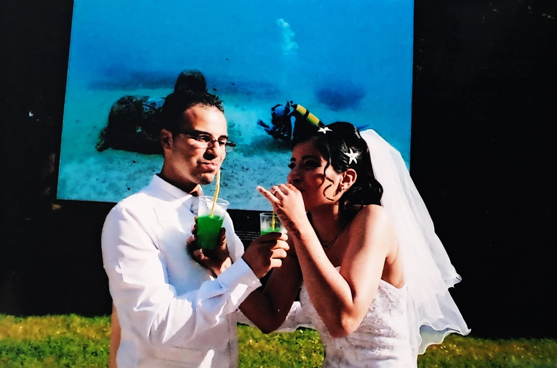 Un couple habillé en mariés, buvant des cocktails verts, avec un tableau sous l'eau en arrière-plan.