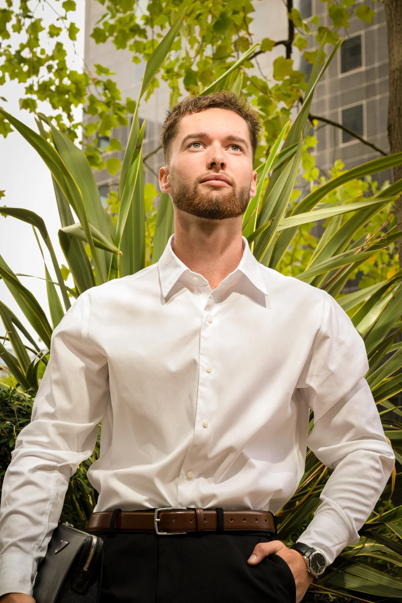 Jeune homme d'affaires en chemise blanche avec un portefeuille, posé devant des plantes urbaines et un bâtiment