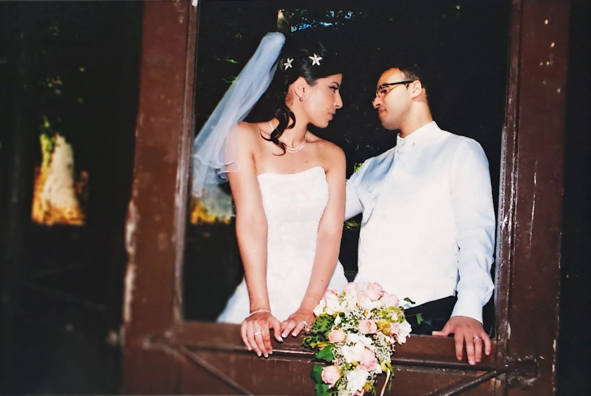 Un couple en mariage regarde tendrement dans les yeux, vu à travers une fenêtre en bois, avec un bouquet de fleurs roses posé sur le rebord.
