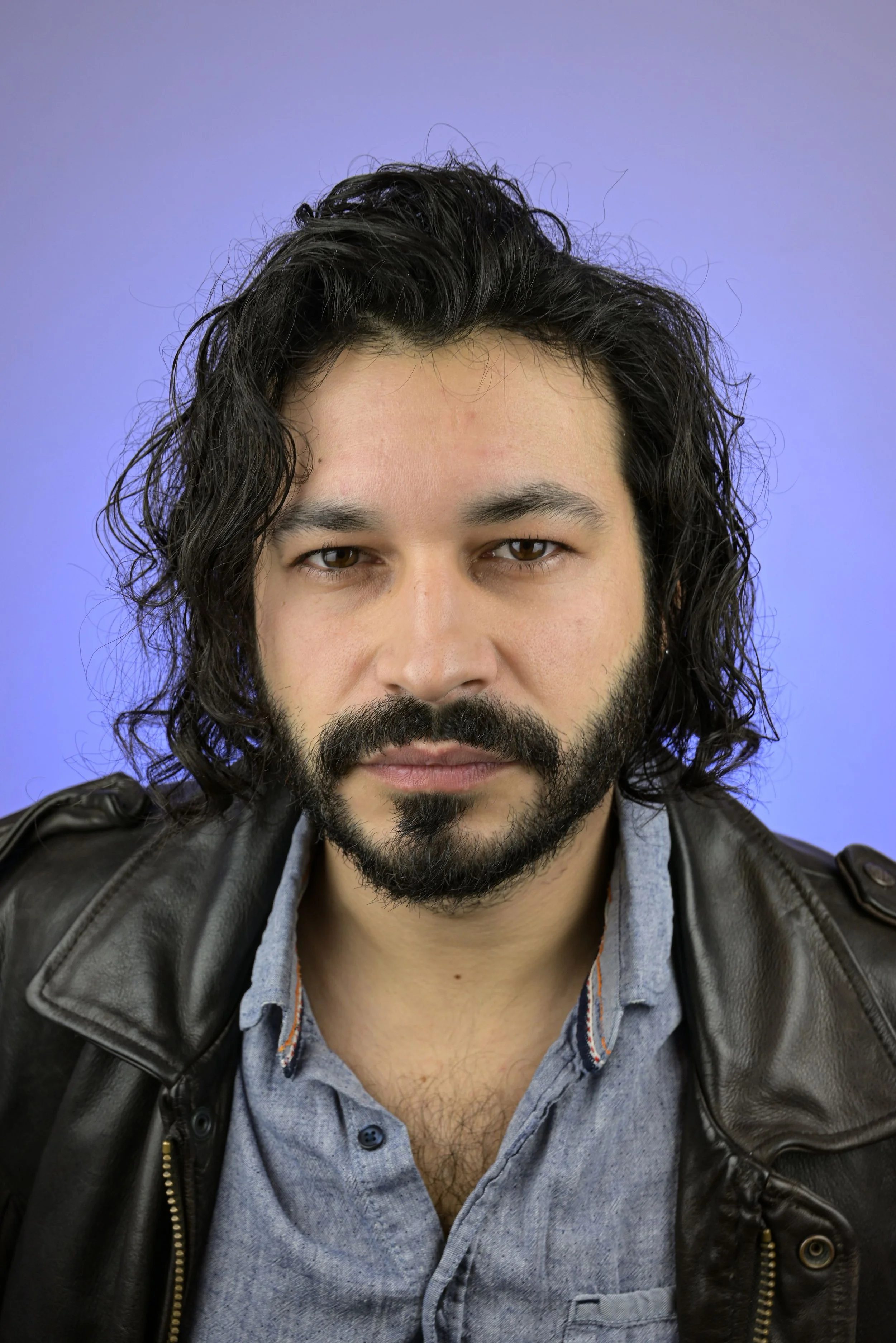 Portrait d'un acteur avec des cheveux longs et bouclés, barbe noire, portant une veste en cuir sombre et une chemise grise, devant un fond violet.