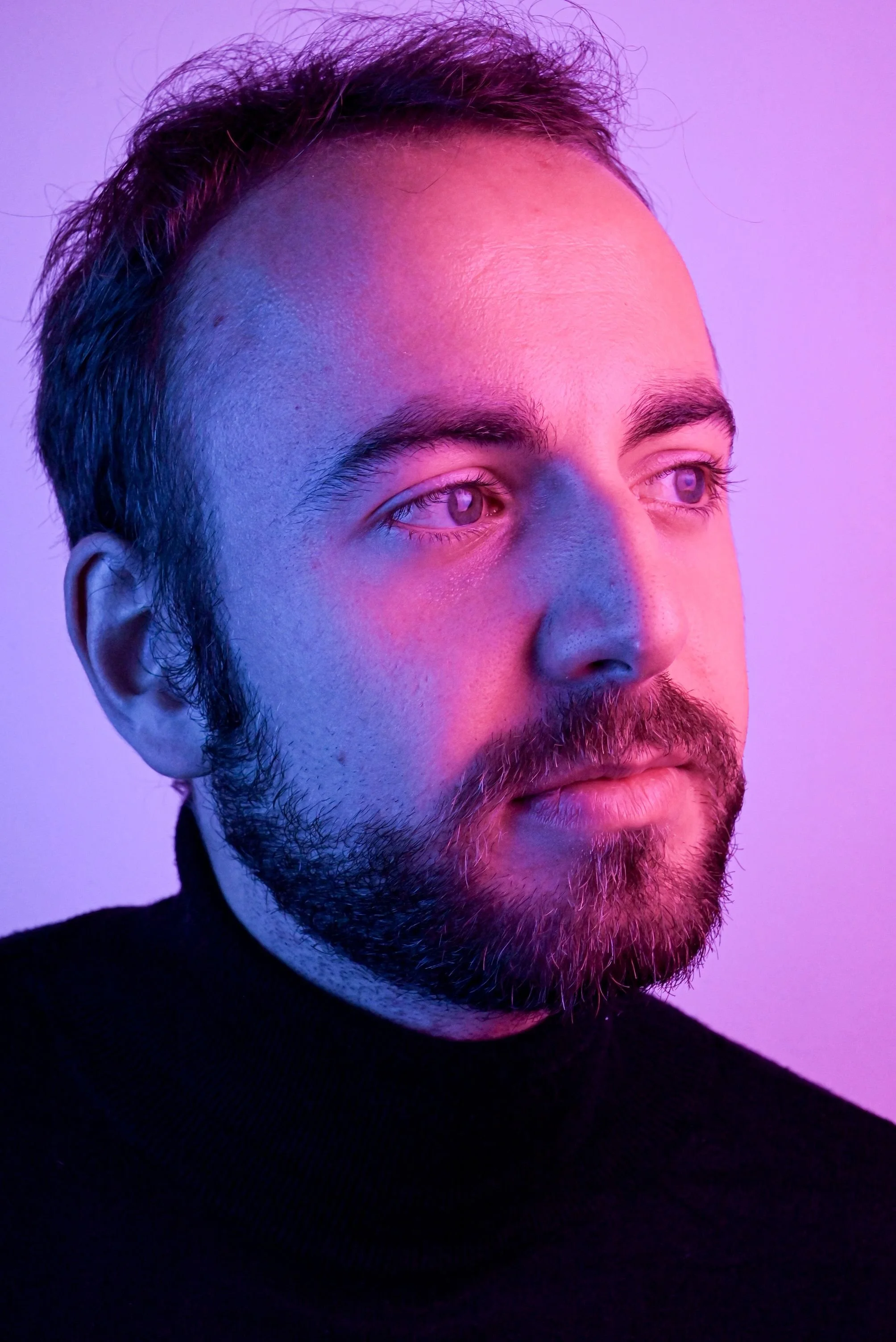 Portrait d'un acteur Thomas Kindler avec une barbe, regardant vers la droite, sous un éclairage violet et rose.