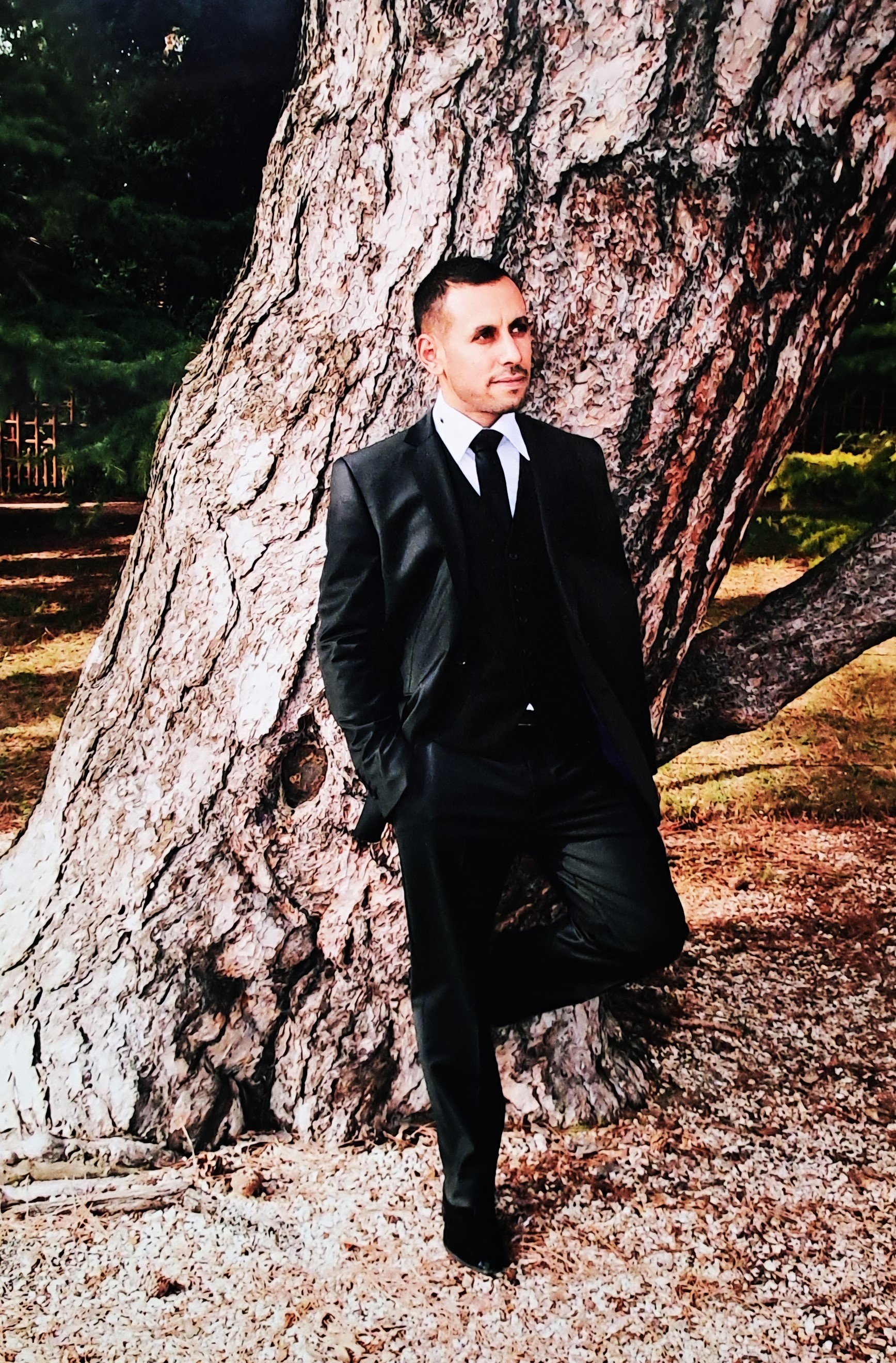 Un homme habillé en costume noir, debout devant un arbre, dans un parc.
