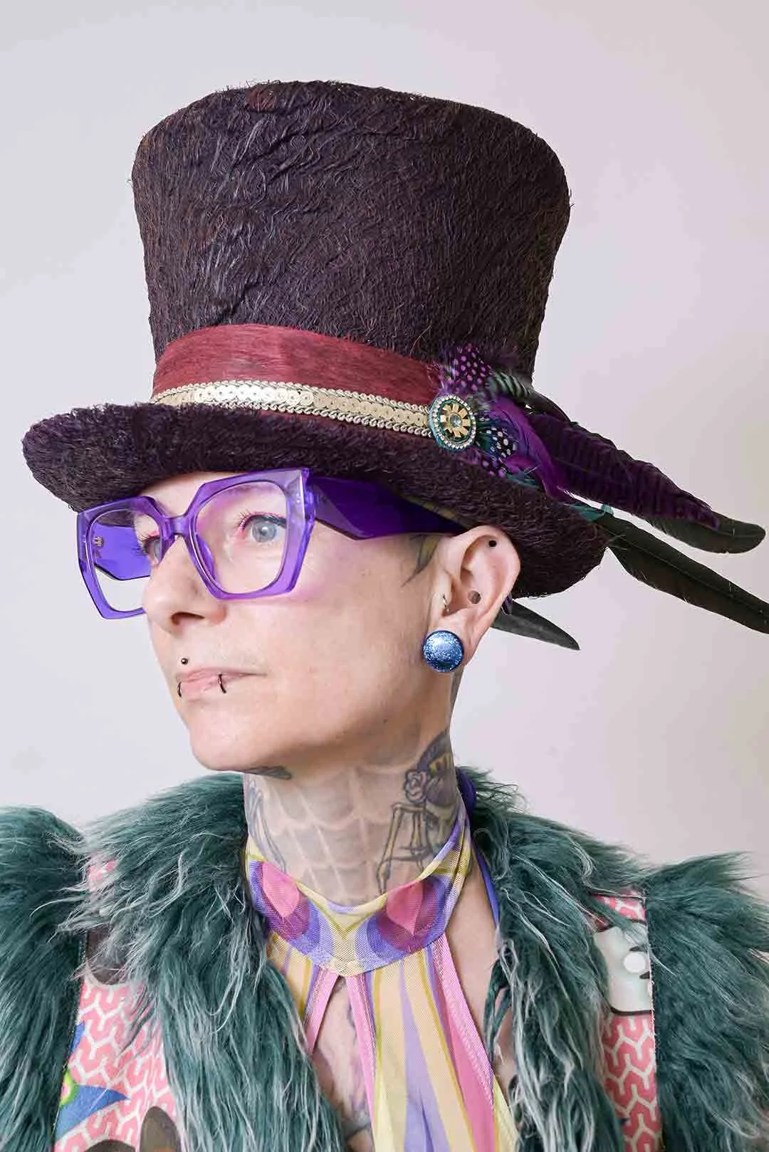 Portrait d'une artiste portant un chapeau haut de forme orné de plumes, de lunettes violettes, et habillée de vêtements colorés avec des tatouages visibles sur le cou.