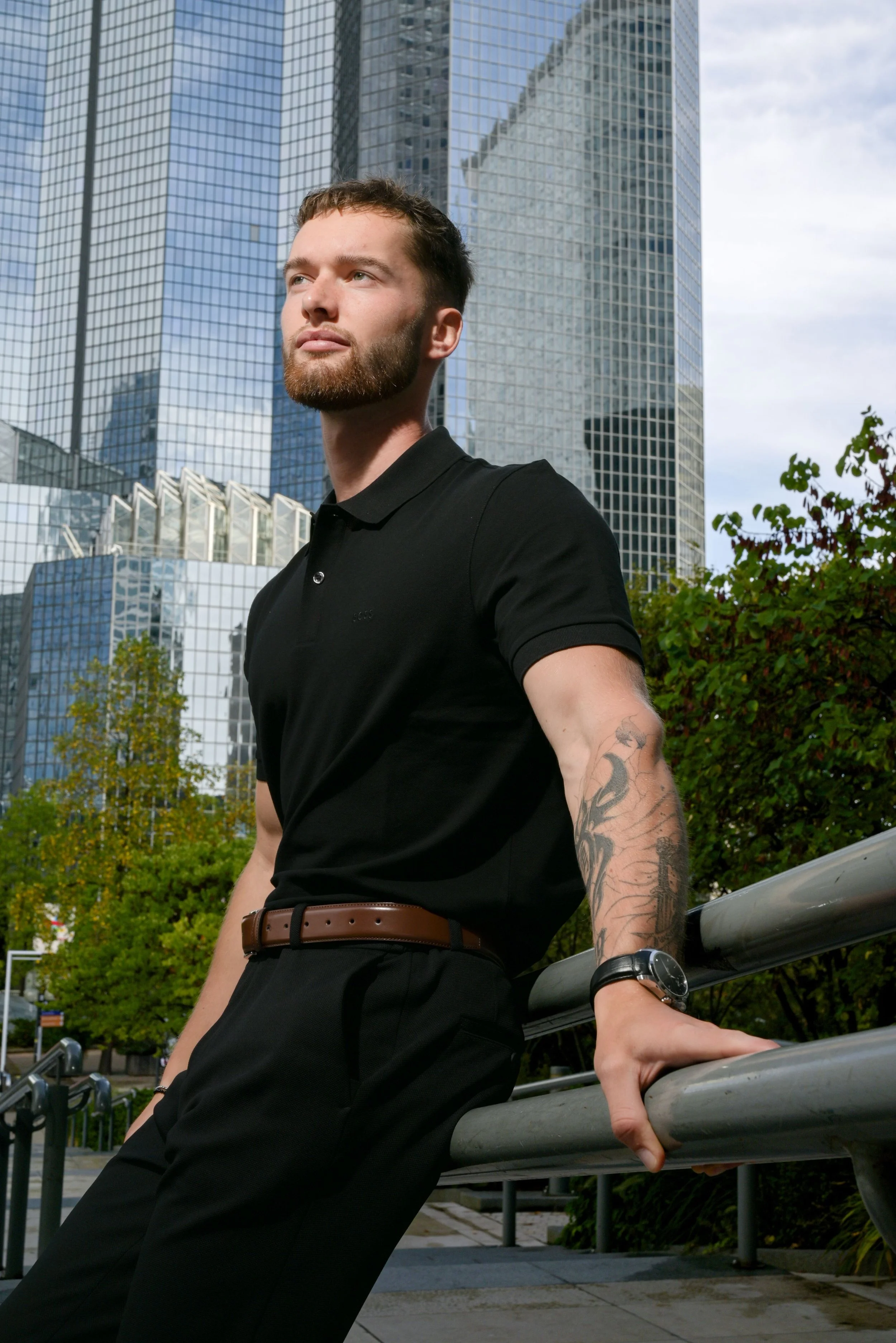 Un homme avec une barbe, portant un polo noir, des pantalons noirs, une ceinture marron, une montre noire, et un tatouage sur le bras gauche, se tient près d'une barrière en métal à l'extérieur avec des gratte-ciel modernes en arrière-plan, sous un c