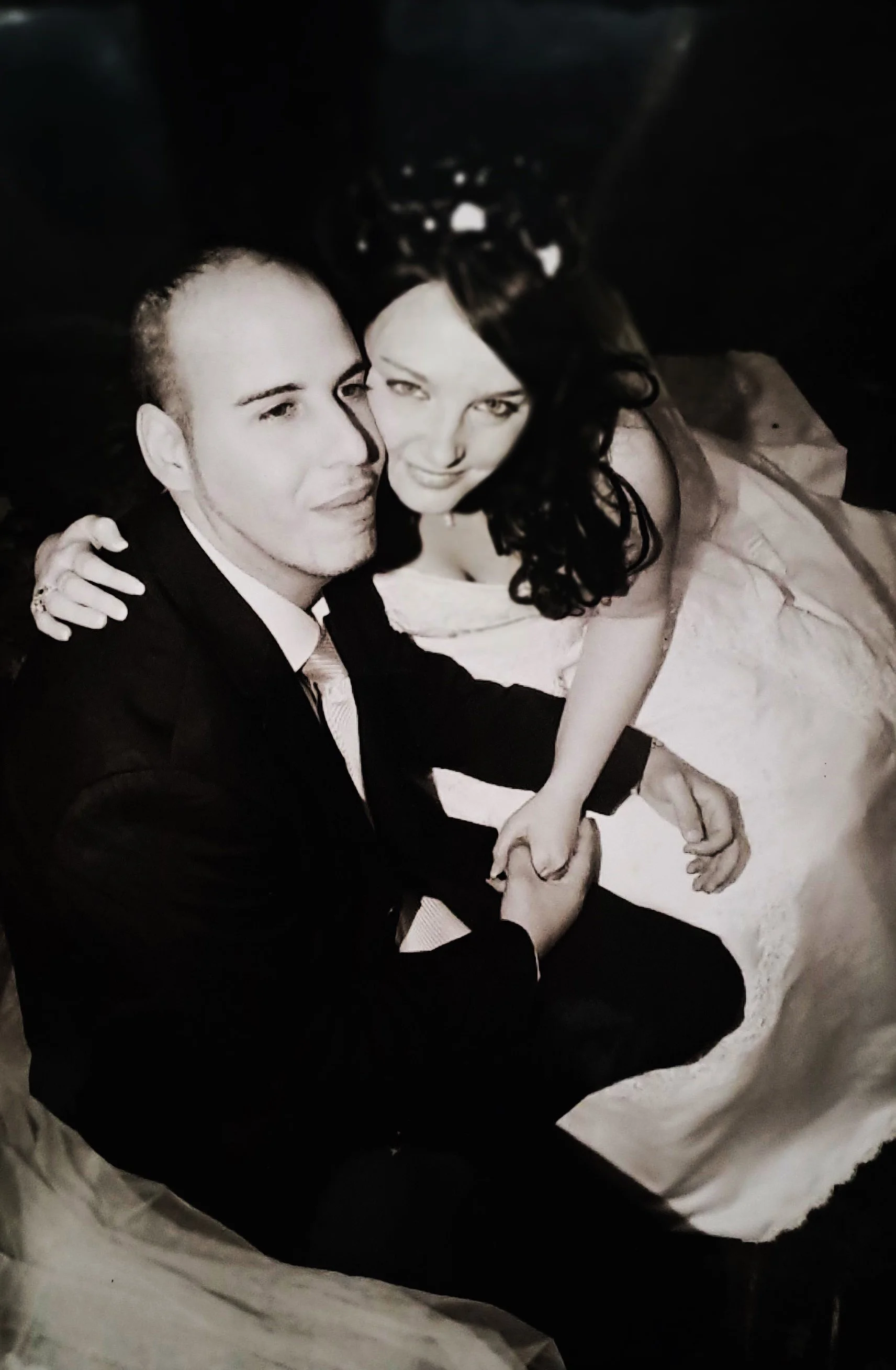 Un couple en costume de mariage, assis et se tenant par la main, dans une photo en noir et blanc.