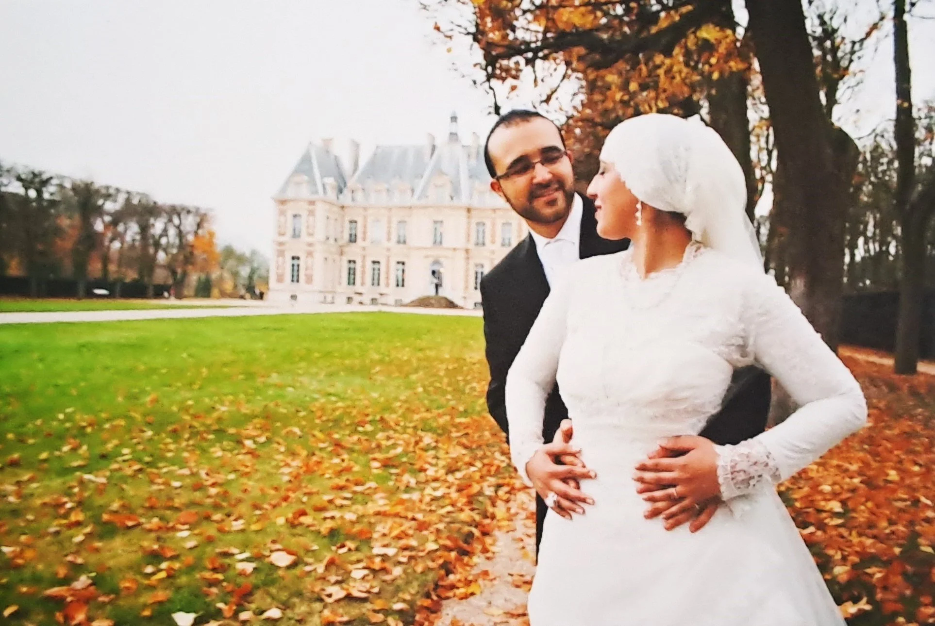 Un couple de mariés pose devant un château en automne, entouré d'arbres aux feuilles orange et rouge, et de feuilles tombées au sol.