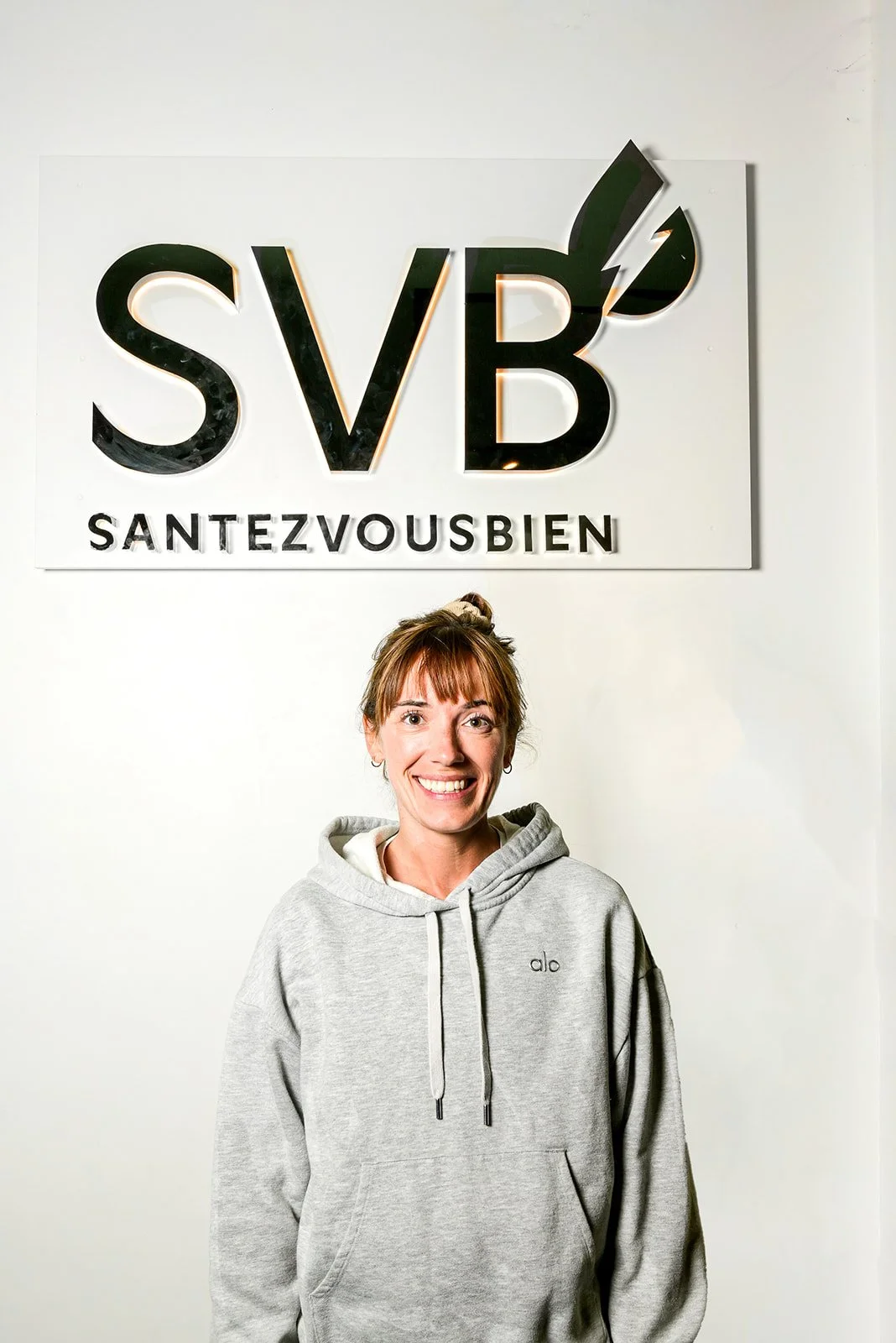 Séance photo portrait corporate d'une femme avec de longs cheveux noirs, souriante, portant une vestUne femme souriante portant un sweat gris dans un intérieur, devant un panneau avec le logo 'SVB' et la phrase 'SANTEZVOUSBIEN'.